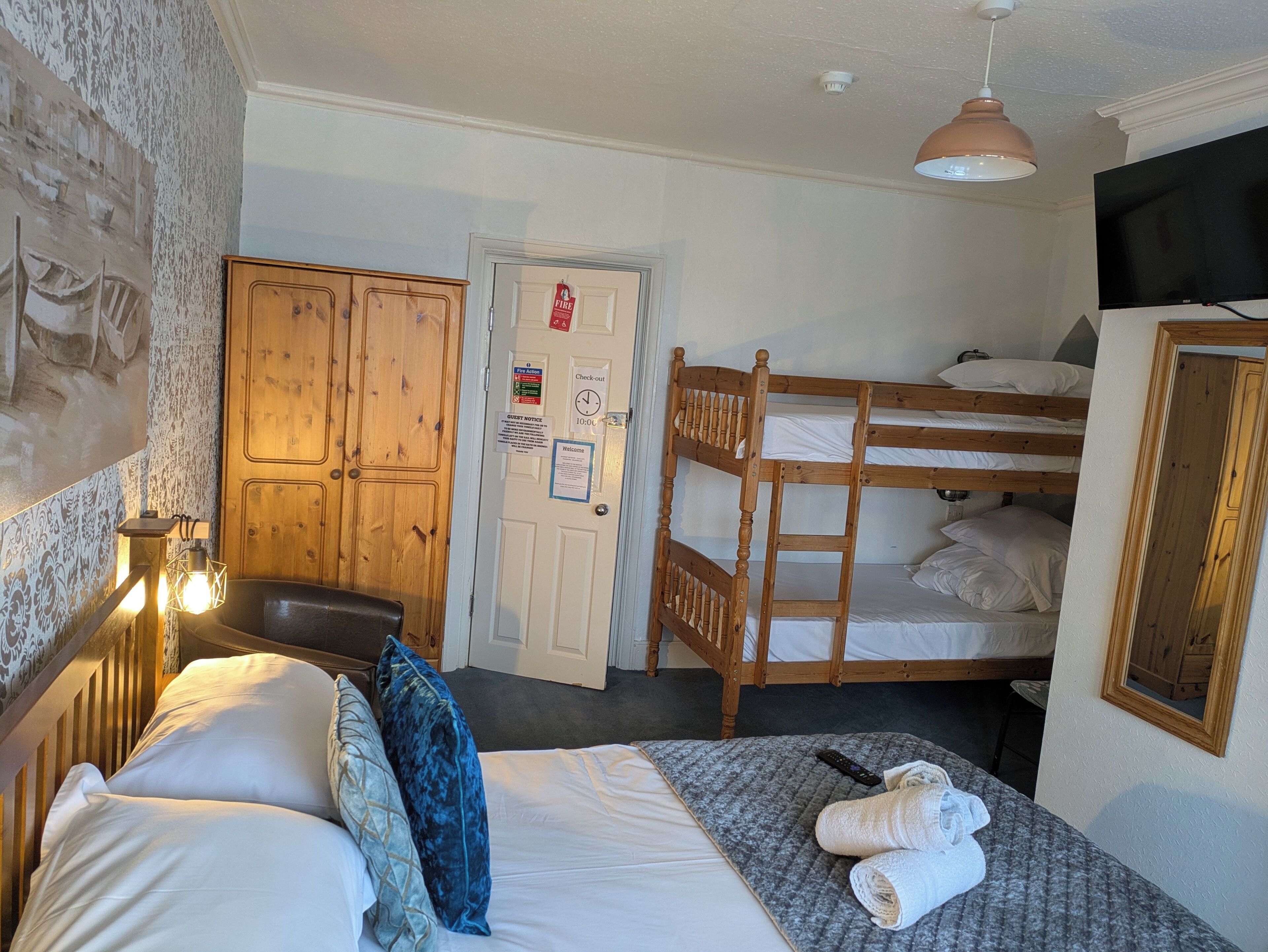Quarto com cama, beliche, guarda-roupa de madeira, escrivaninha com luminária e televisão fixada na parede.