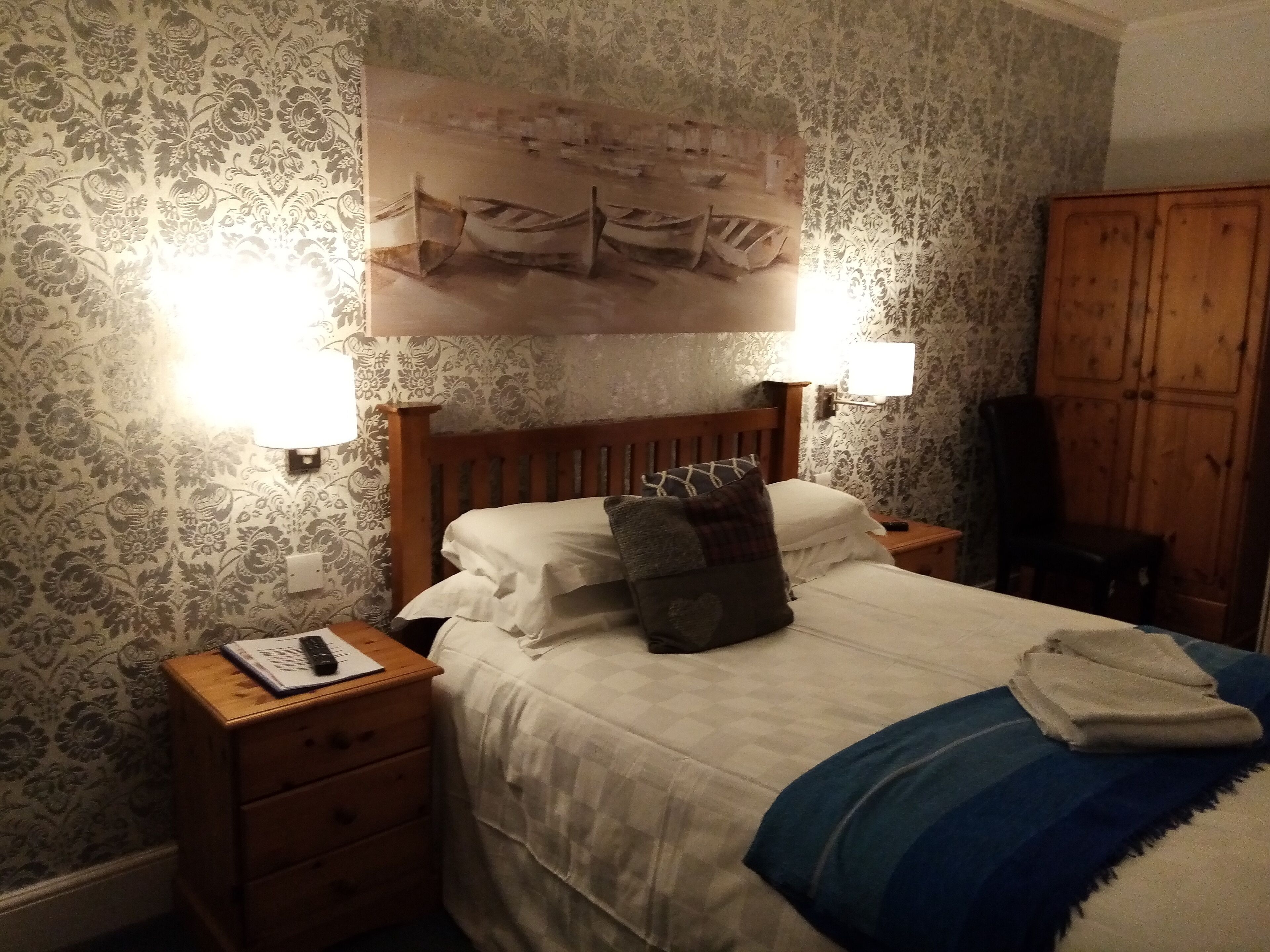 A bedroom with a wooden headboard, two bedside lamps, a bed with white bedding and a blue blanket, a wooden nightstand with a book and remote, and a patterned wallpaper.