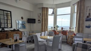 A room with a large window offering a view of the sea, a television mounted on the wall, and a variety of seating arrangements including tables and chairs. - The Kenton (Scarborough)