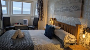 A hotel room with a bed, a desk, a chair, a window with a view of the sea, and a painting on the wall. - The Kenton (Scarborough)