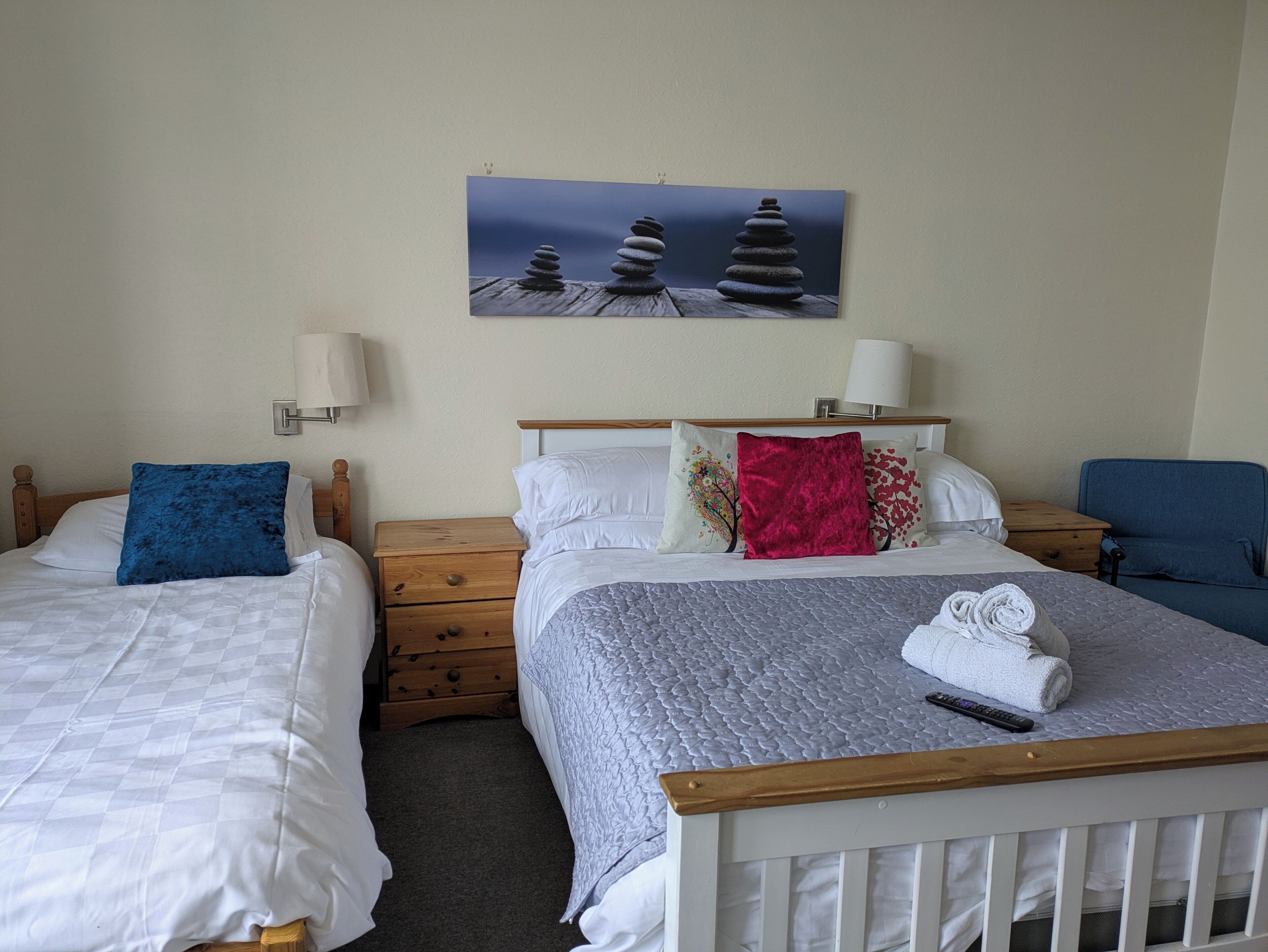 A hotel room with two beds, a wooden headboard, a nightstand, a lamp, a painting, and a blue chair.