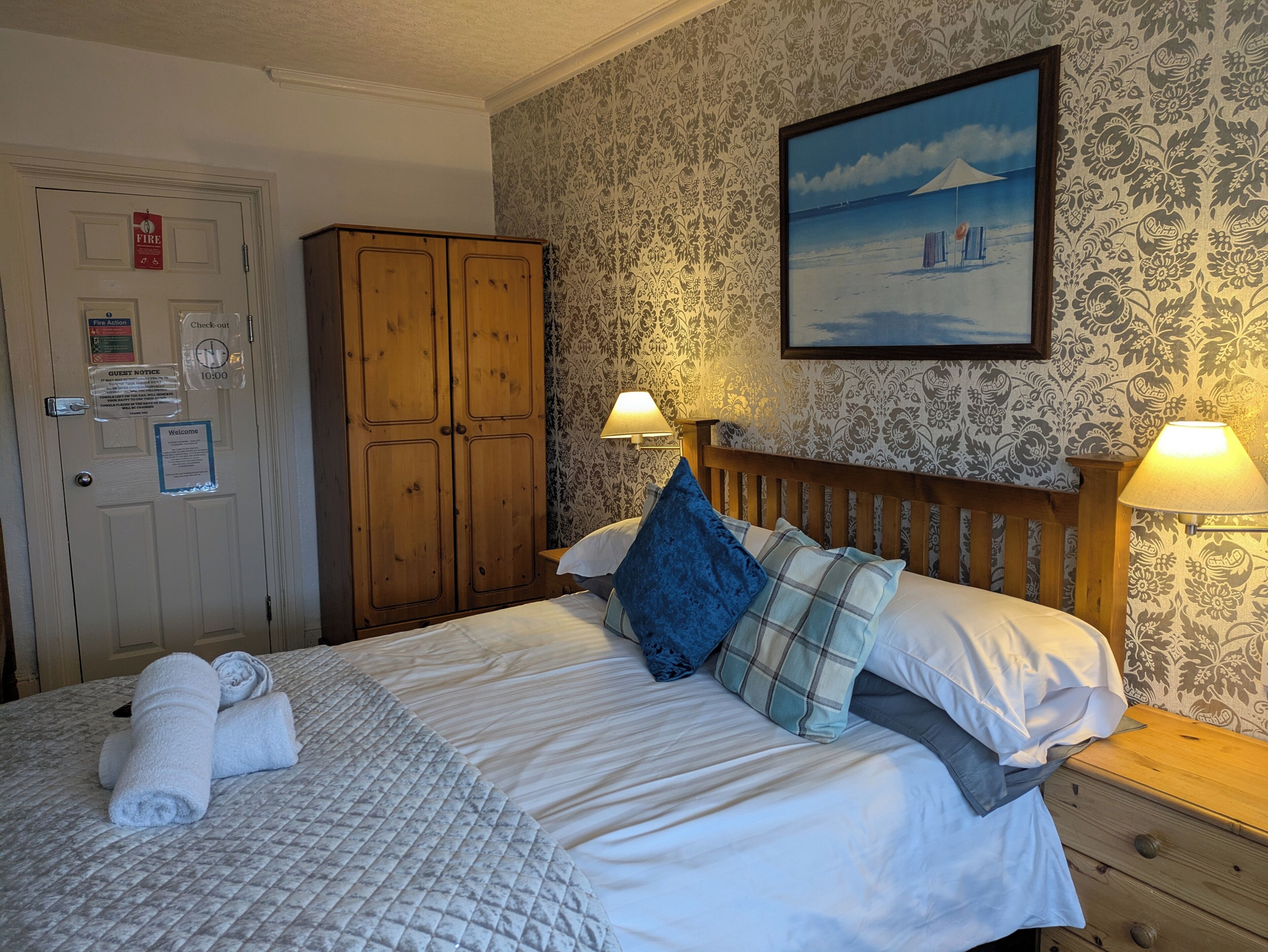 A hotel room with a bed, a wooden wardrobe, a painting on the wall, and a fire exit sign.