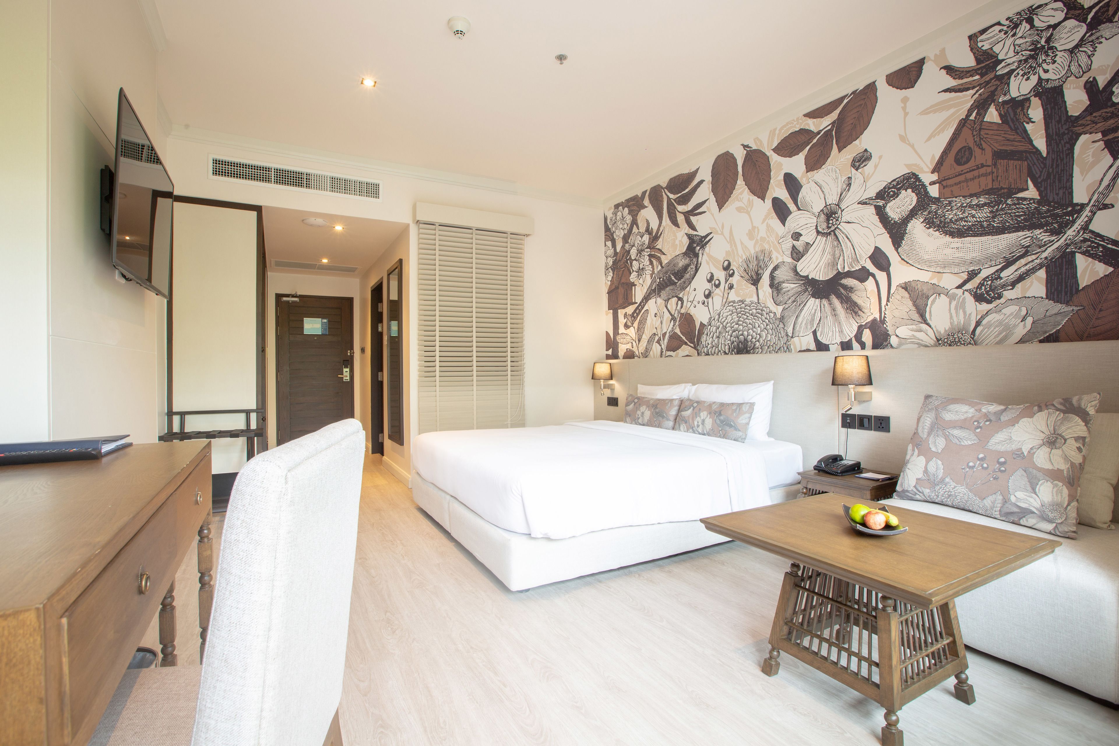 Una habitación de hotel con cama, un escritorio con computadora, una mesa de centro con fruta, un sofá y un mural en la pared.