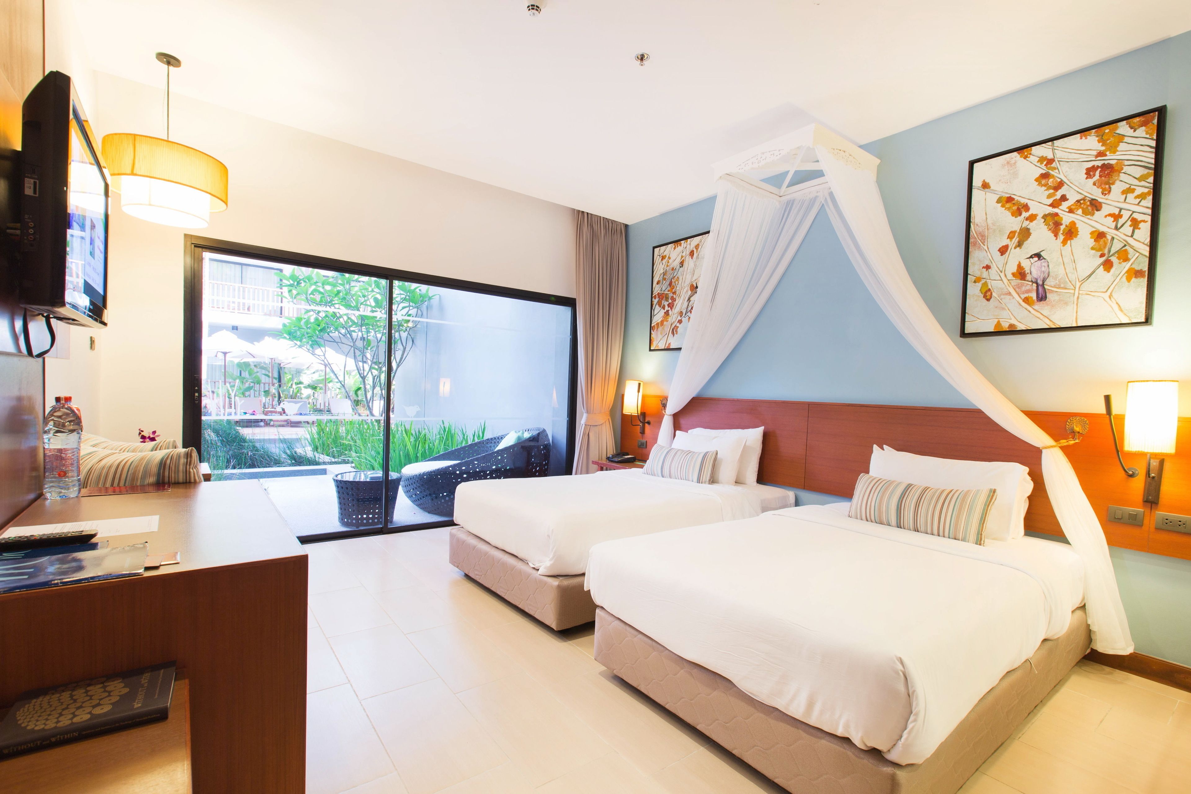 A hotel room with two beds, a desk, a TV, and a large window with a view of greenery.