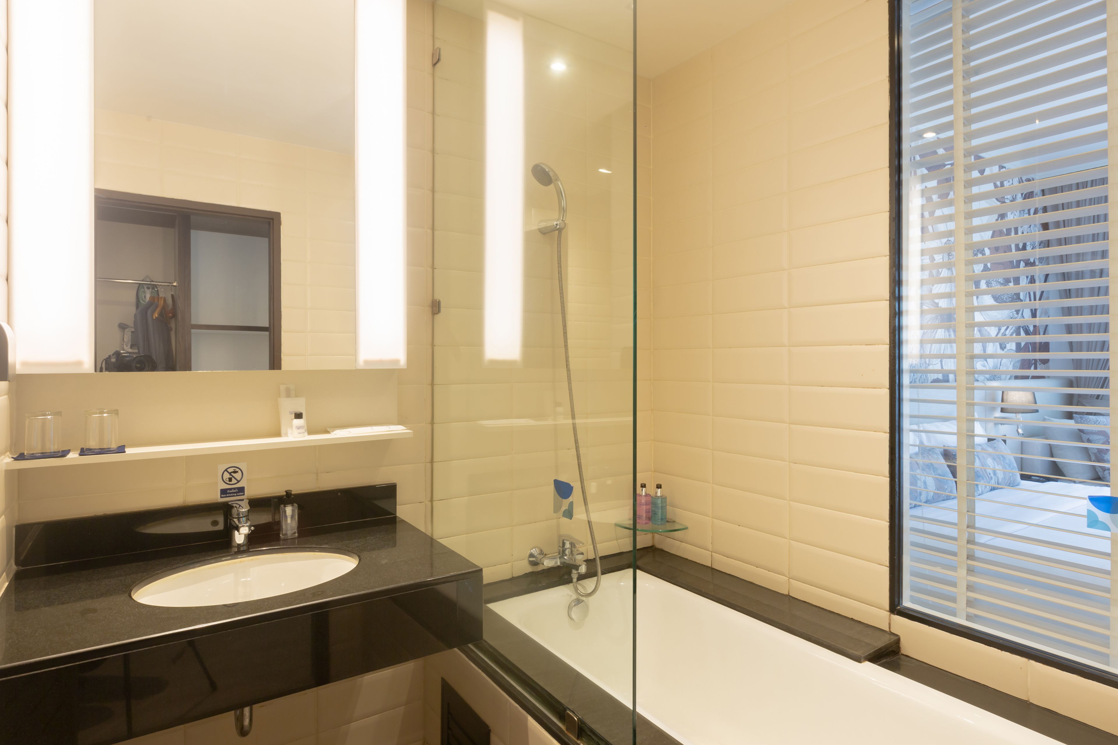 A modern bathroom with a glass-enclosed shower, a white sink, and a large window with blinds.