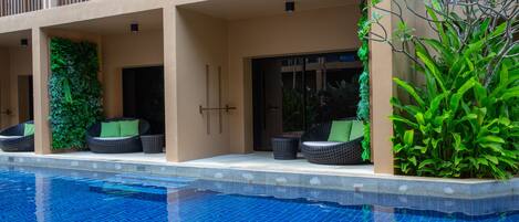 Un área al lado de la piscina con una piscina azul cristalina, mobiliario exterior con cojines verdes y un balcón con barandilla.