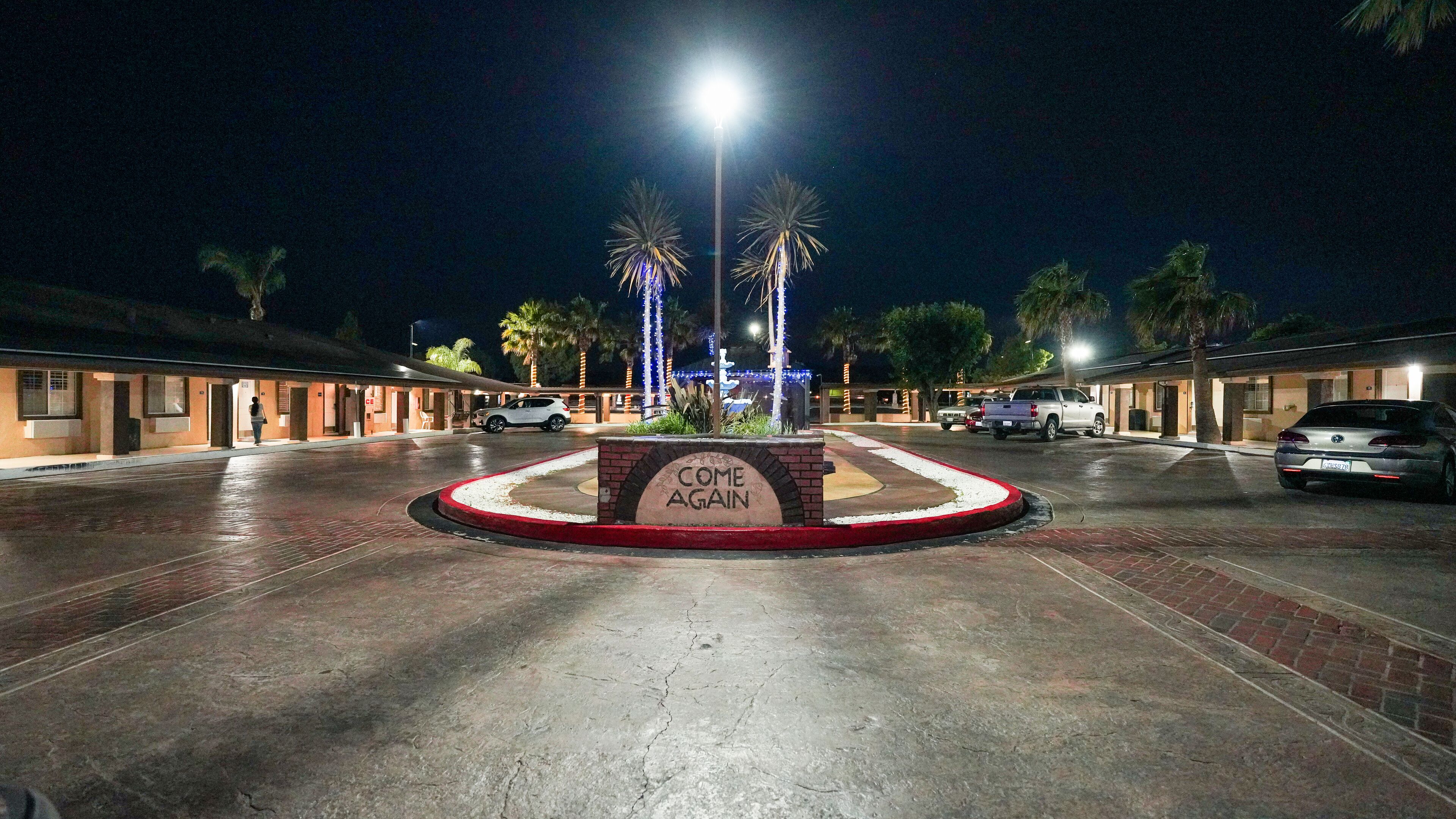 A motel with a 'COME AGAIN' sign in the center, a parking lot with several cars, and palm trees in the background.
