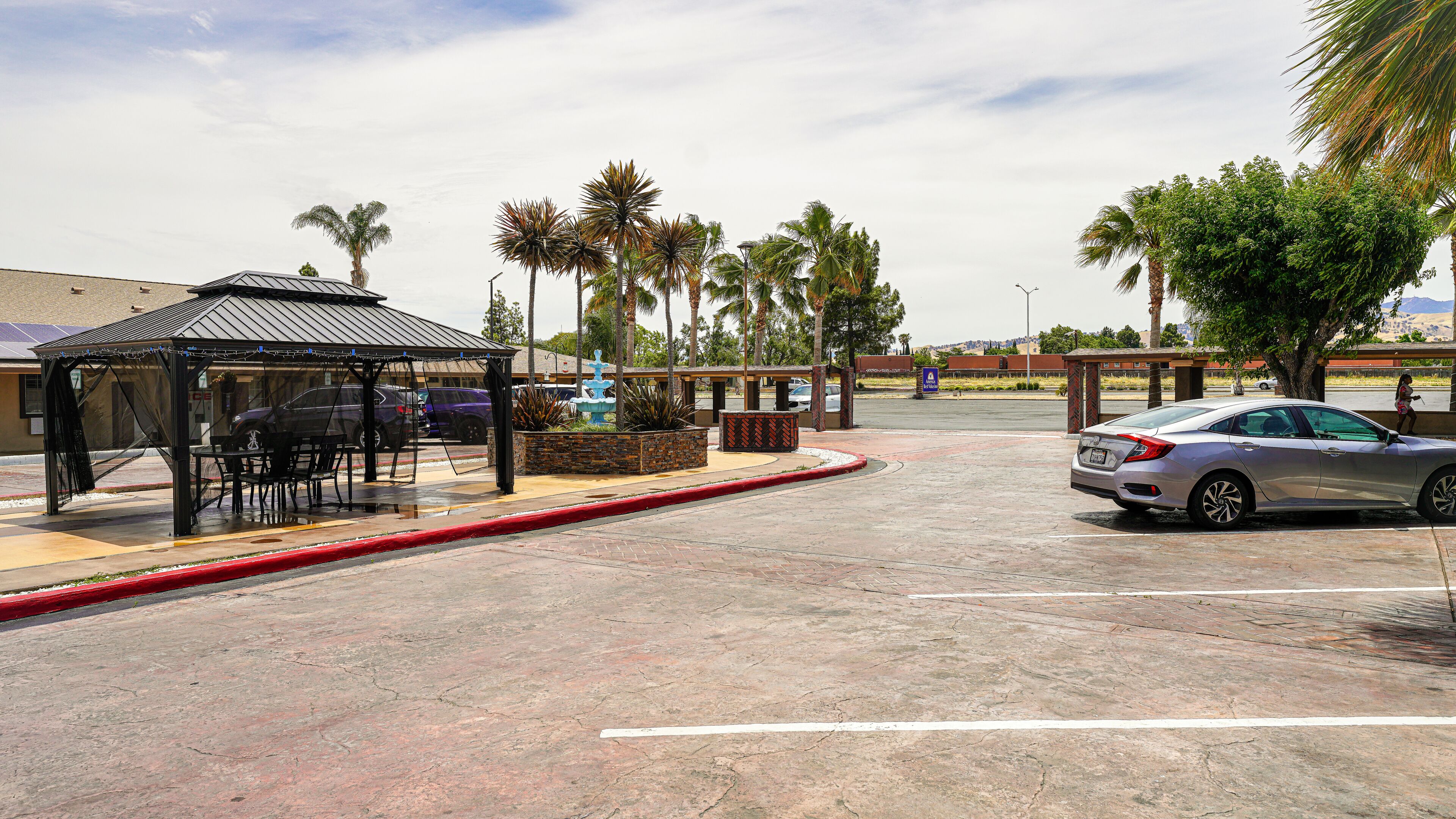 A covered outdoor seating area with a gazebo, a parking lot with cars, and palm trees.