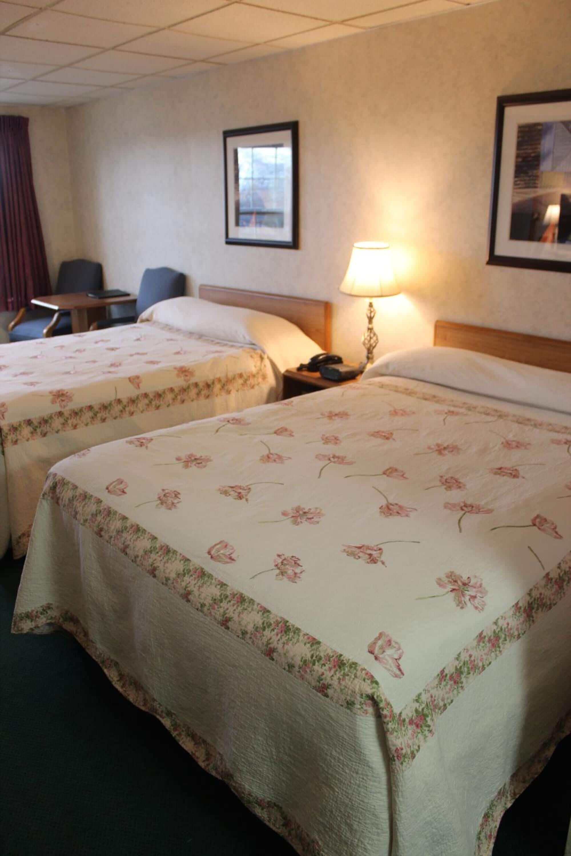 A hotel room with two beds, a desk, a chair, a lamp, and framed pictures on the wall.