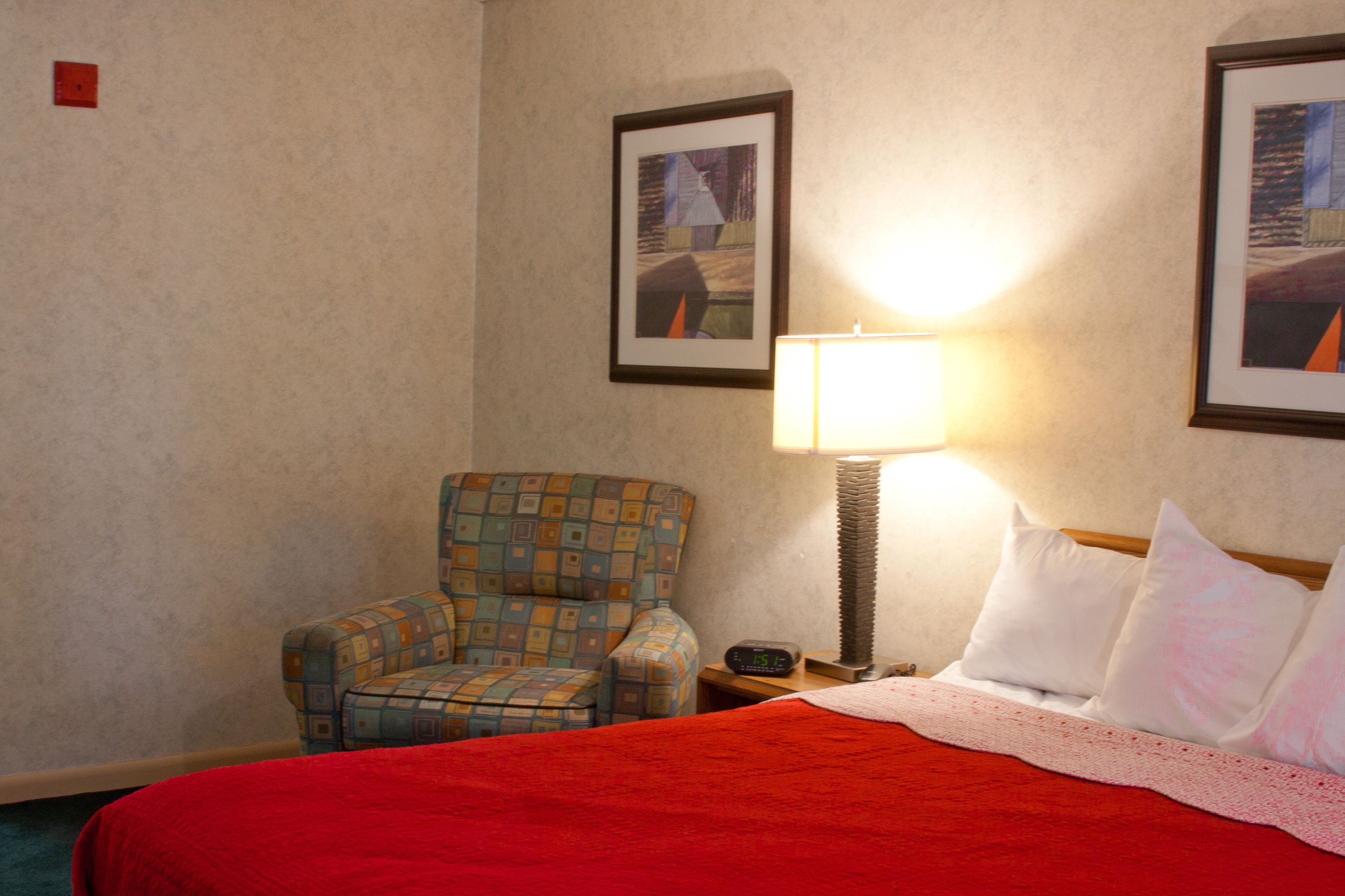 A hotel room with a bed, a chair, a nightstand, a lamp, and two framed pictures on the wall.