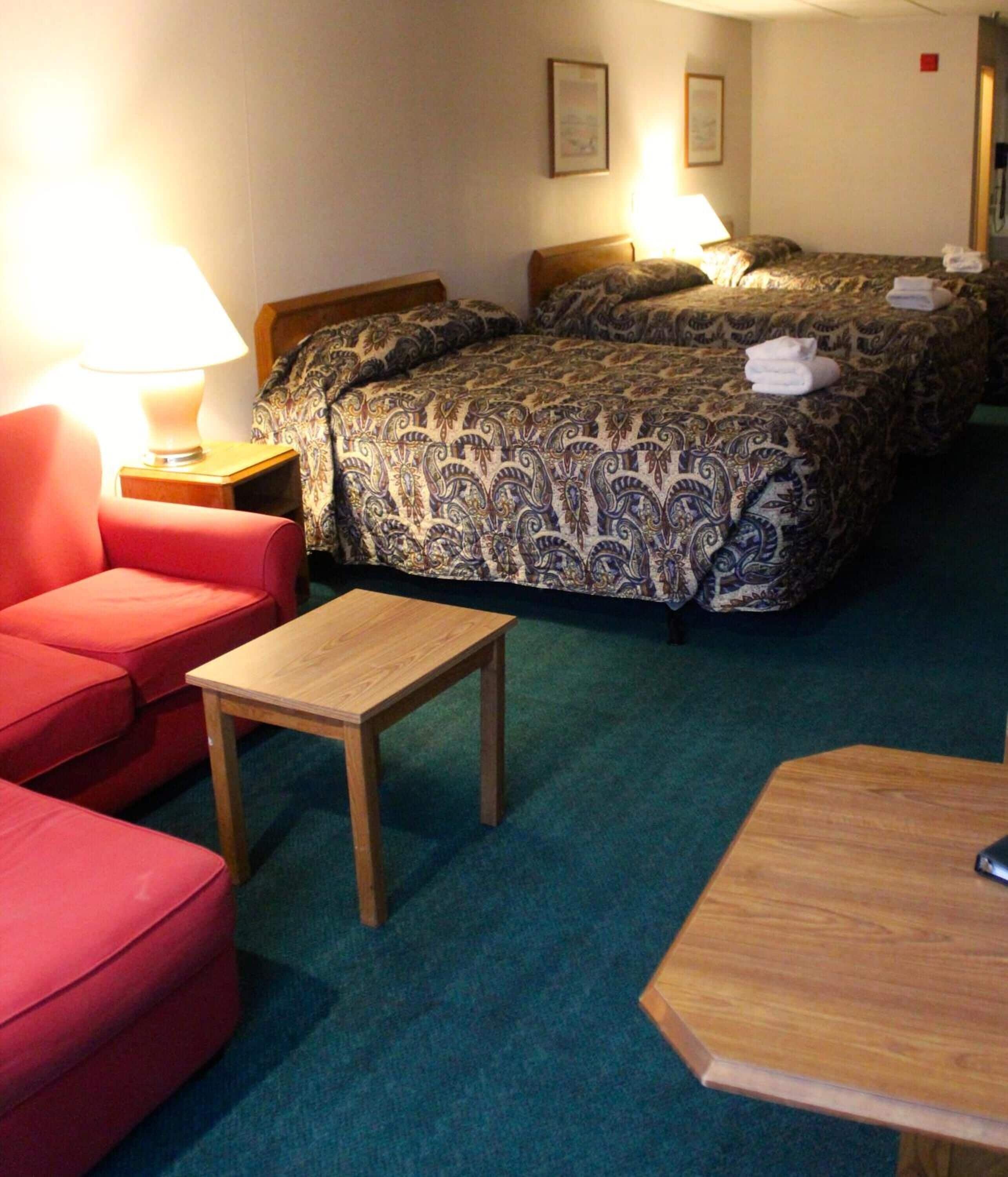 Een hotelkamer met twee bedden, een rode fauteuil, een klein houten tafeltje en een lamp.