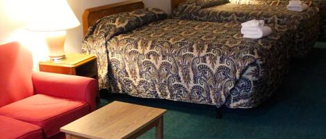 A hotel room with two beds, a red armchair, a small wooden table, and a lamp.