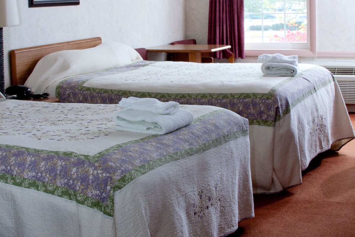 Une chambre d’hôtel avec deux lits, un bureau, une chaise, une fenêtre avec des rideaux et un tableau encadré au mur.