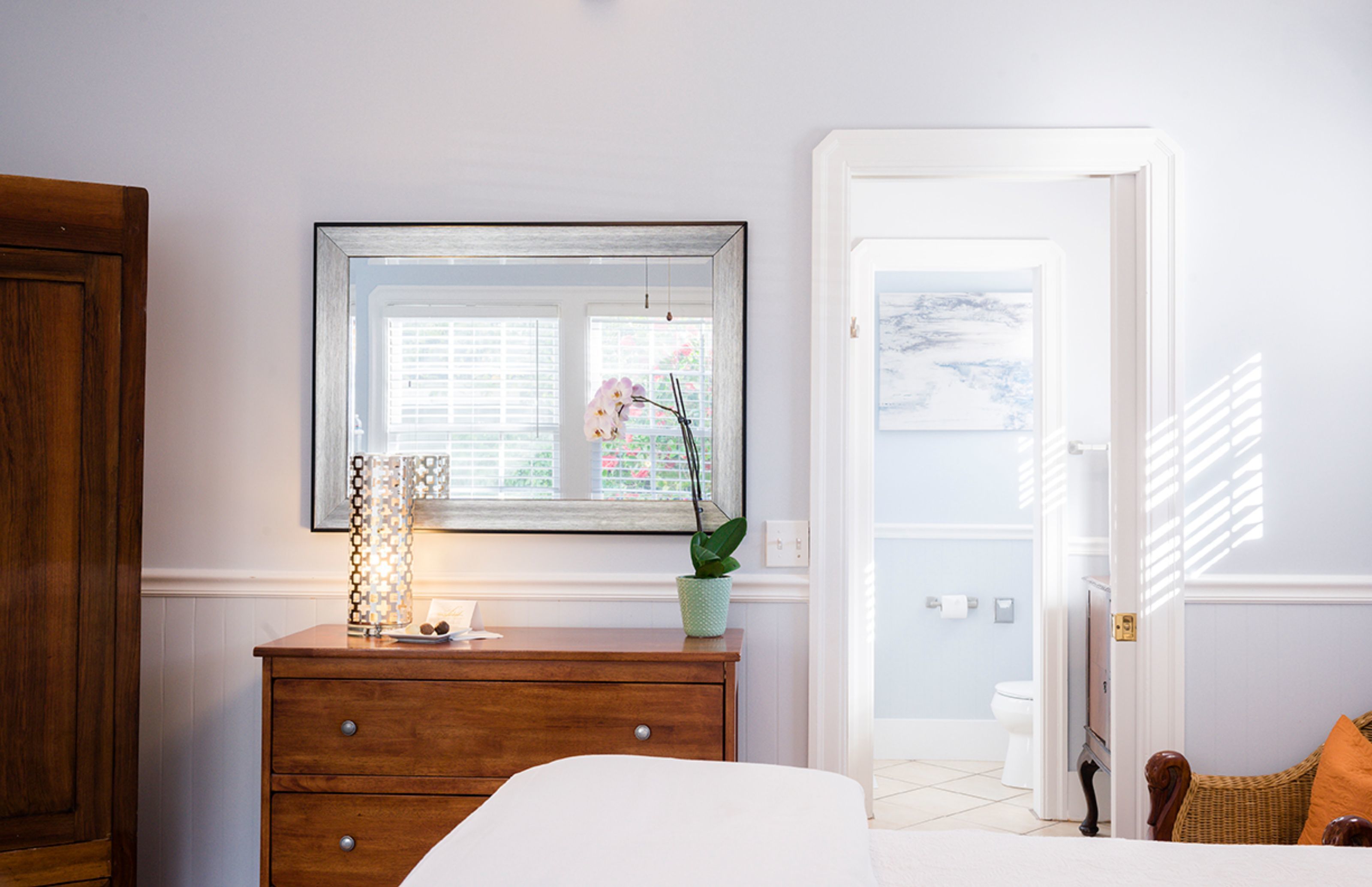 A bedroom with a wooden dresser, a bed, a window with blinds, a potted plant, and a wicker chair.