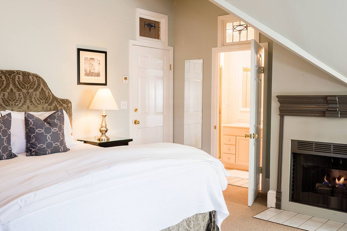 A bedroom with a large bed, a bedside table with a lamp, a framed picture on the wall, and a fireplace.