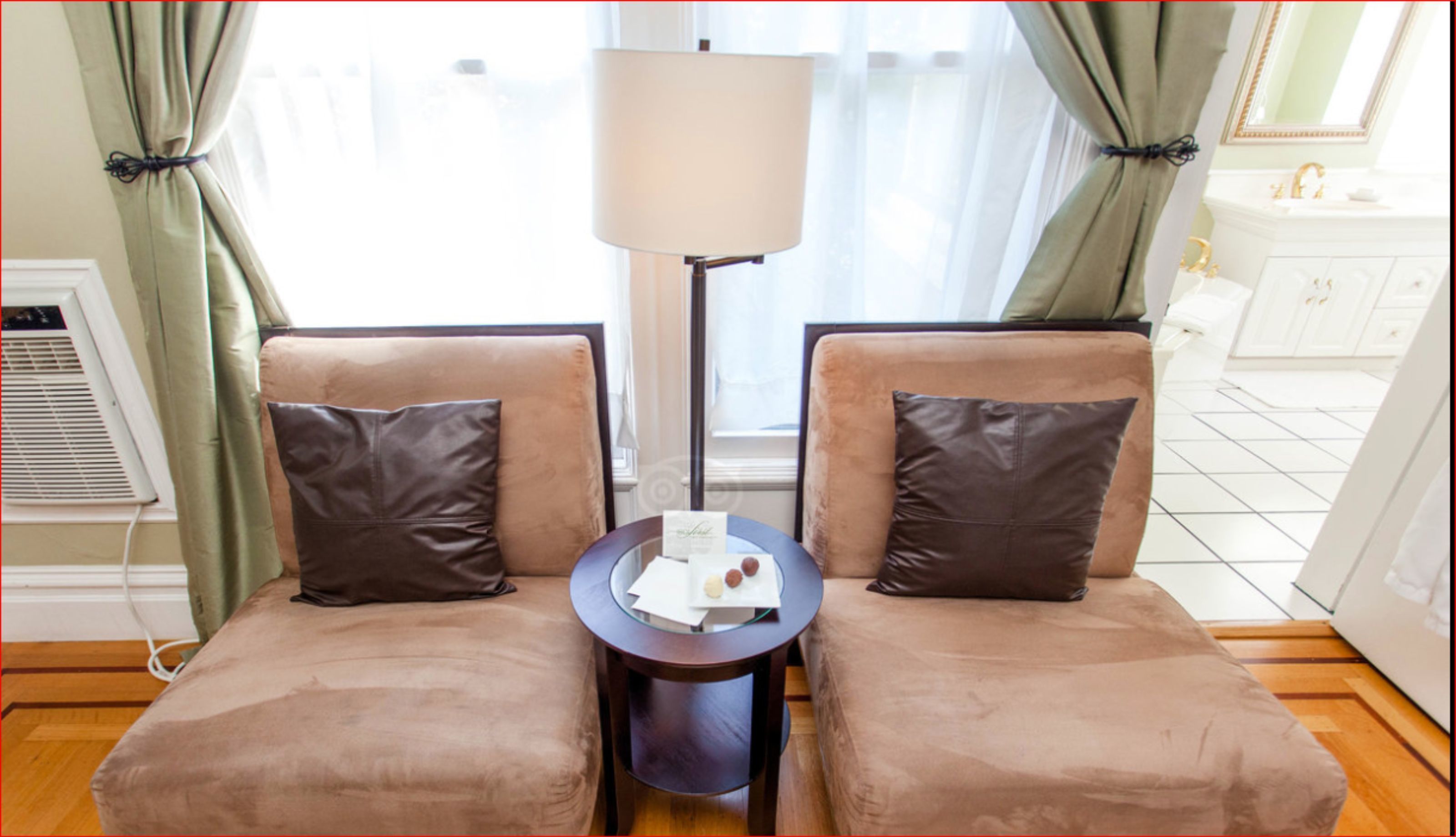 A room with two brown armchairs, a small round table with a lamp, and a view of a bathroom with a white sink and mirror.