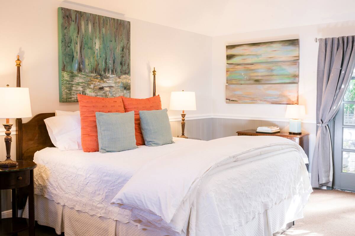 A neatly made bed with white linens and multiple pillows, a nightstand with a lamp, and two paintings on the wall.
