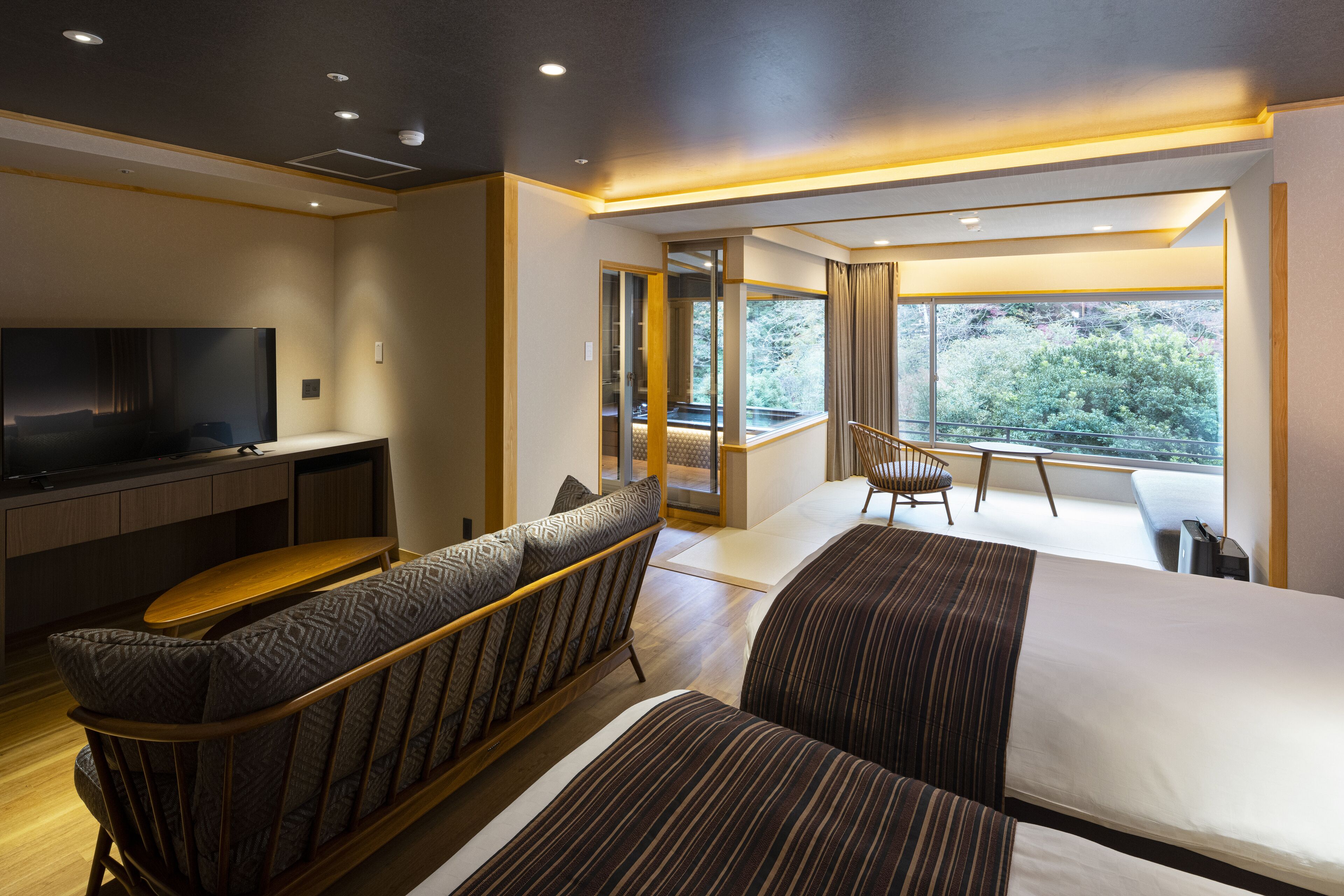 A modern hotel room with a large bed, a sofa, a TV, and a balcony with a view of greenery.