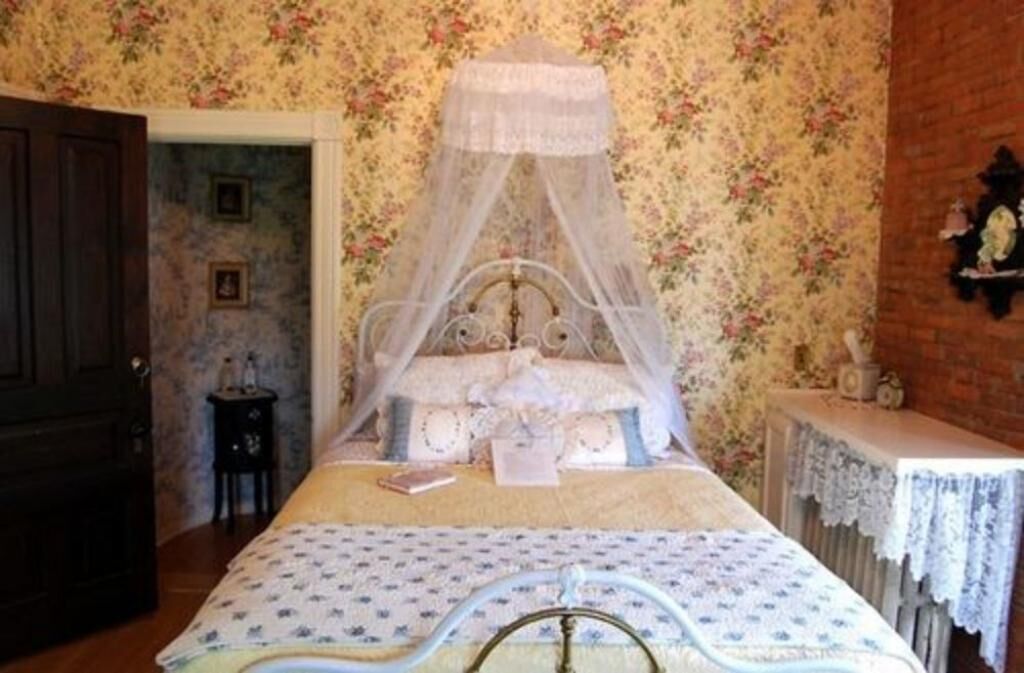 A bedroom with a canopy bed, floral wallpaper, a wooden door, and a small bedside table with a lamp.