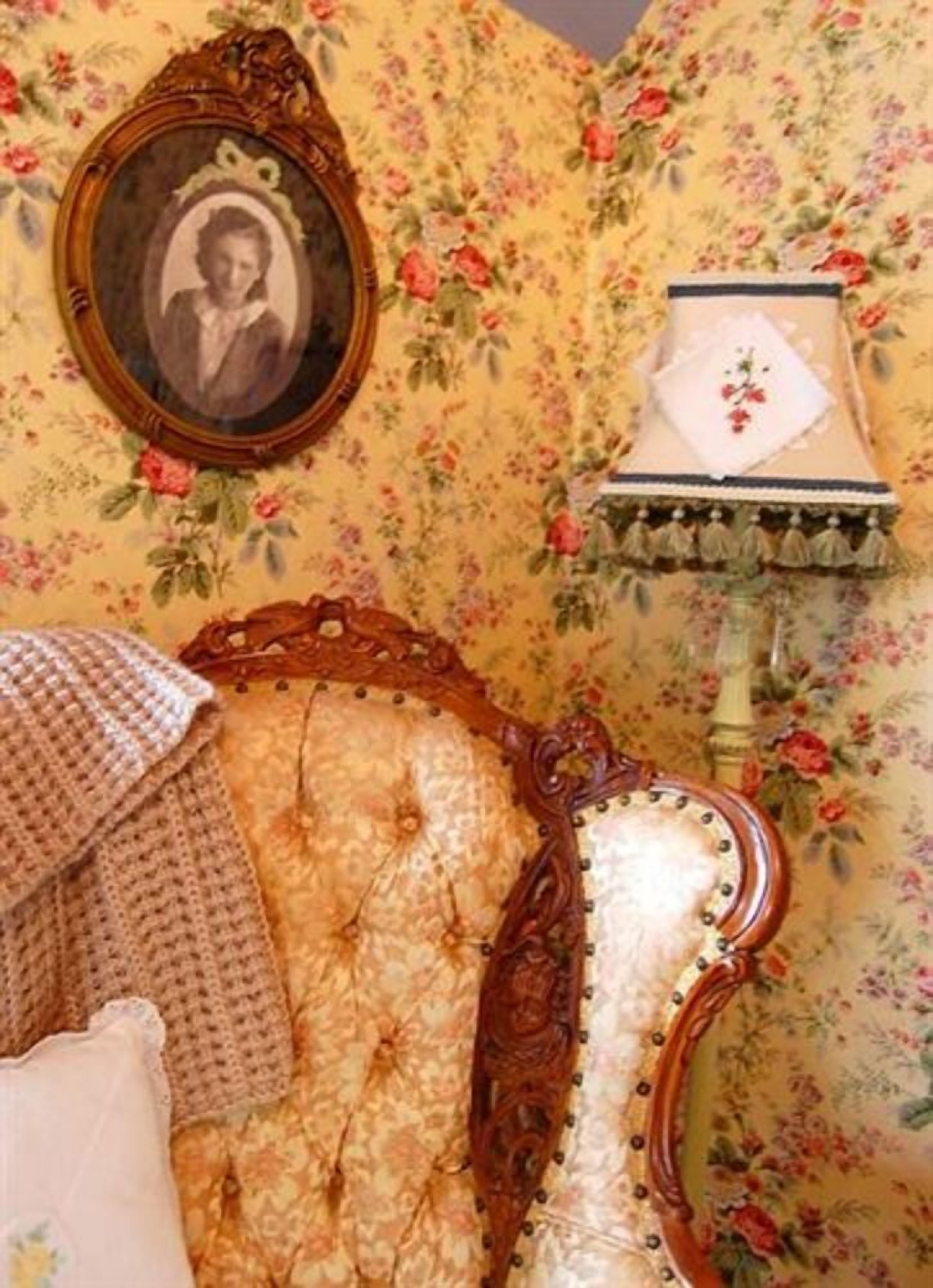 A vintage room with floral wallpaper, a wooden-framed mirror, a decorative lamp, and a knitted blanket.