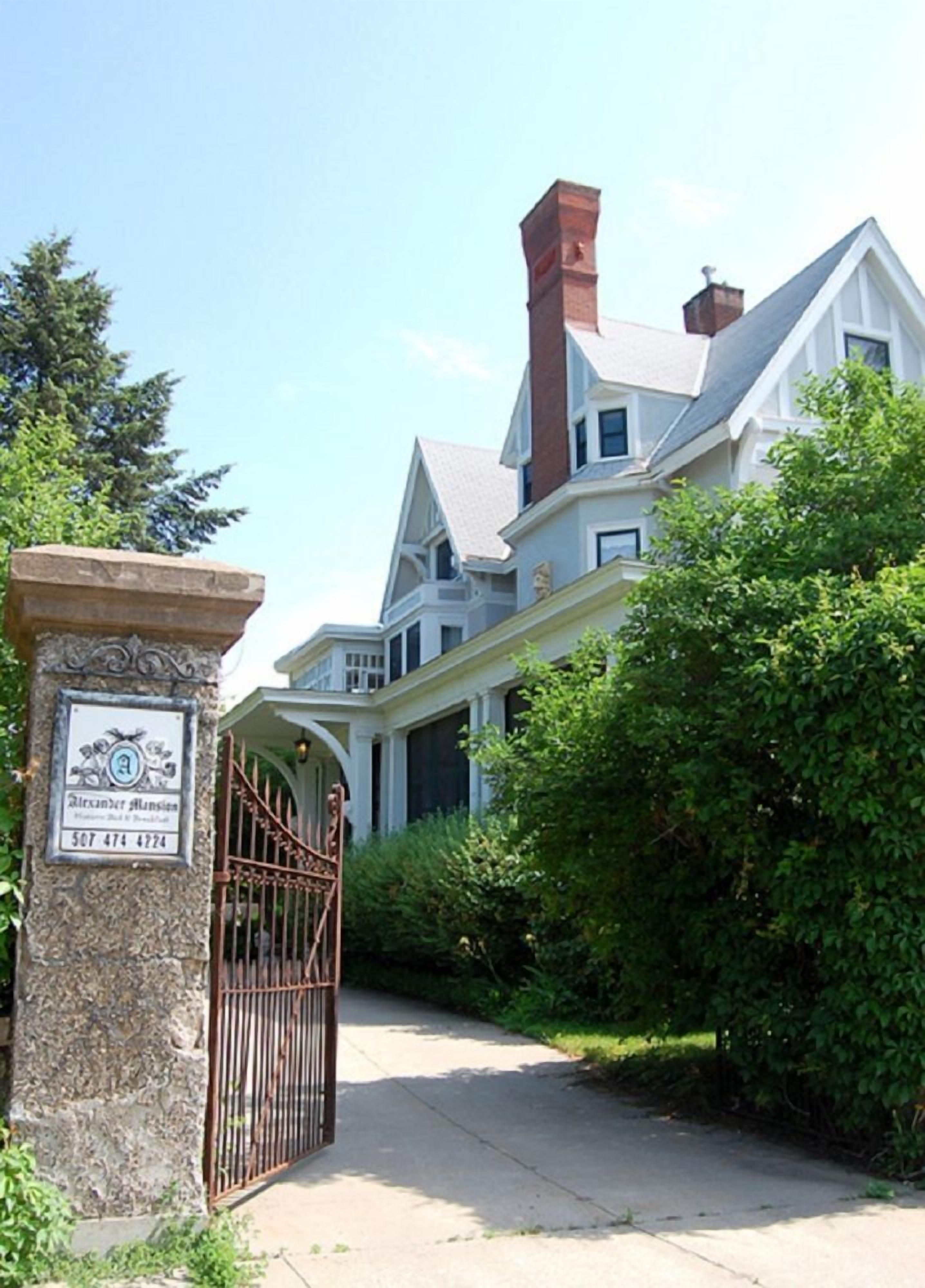 Una casa grande e histórica con una chimenea destacada, una entrada con puerta de rejas y un pilar de piedra con una placa.