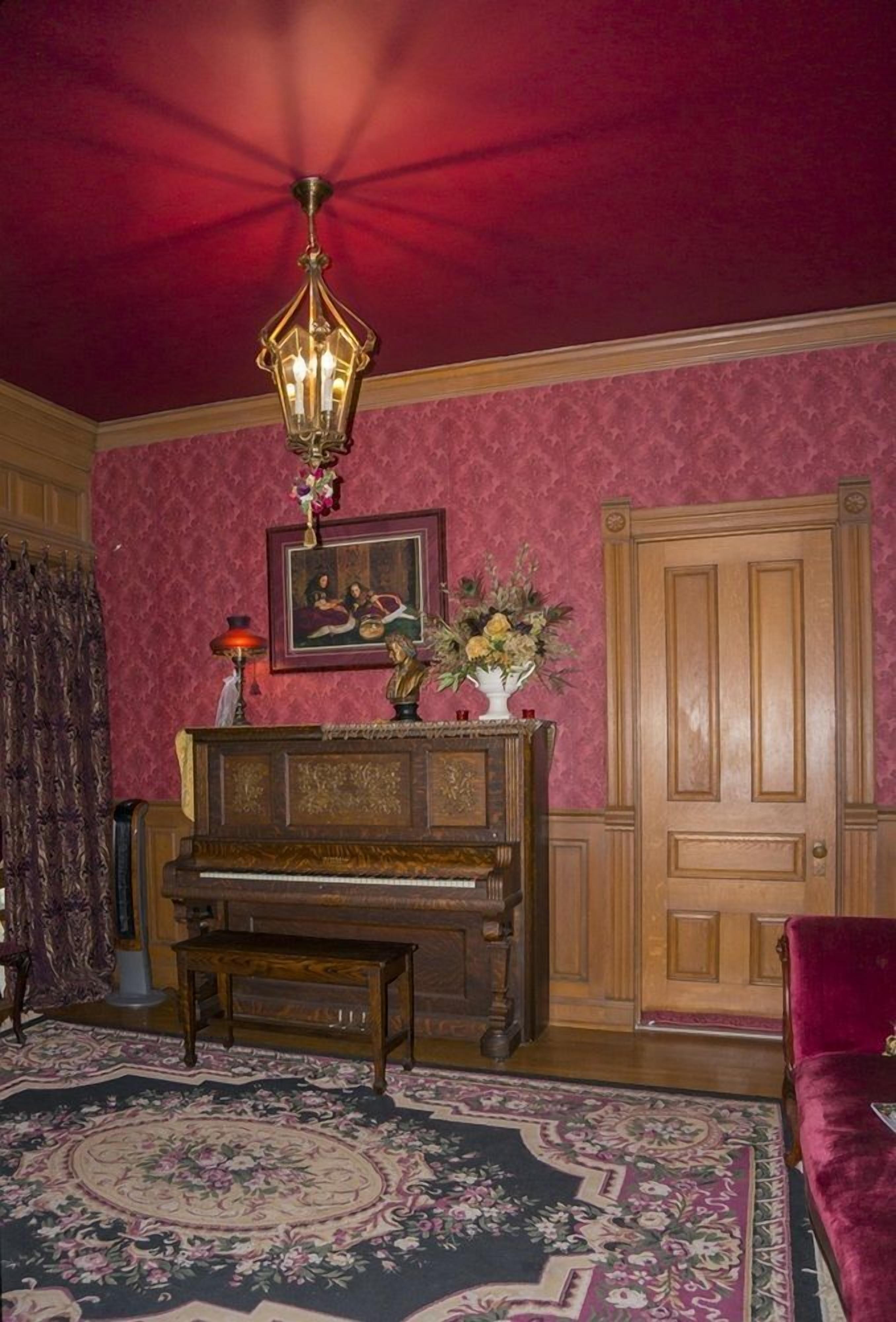 A room with a wooden piano, a red carpet, a wooden door, and a chandelier.