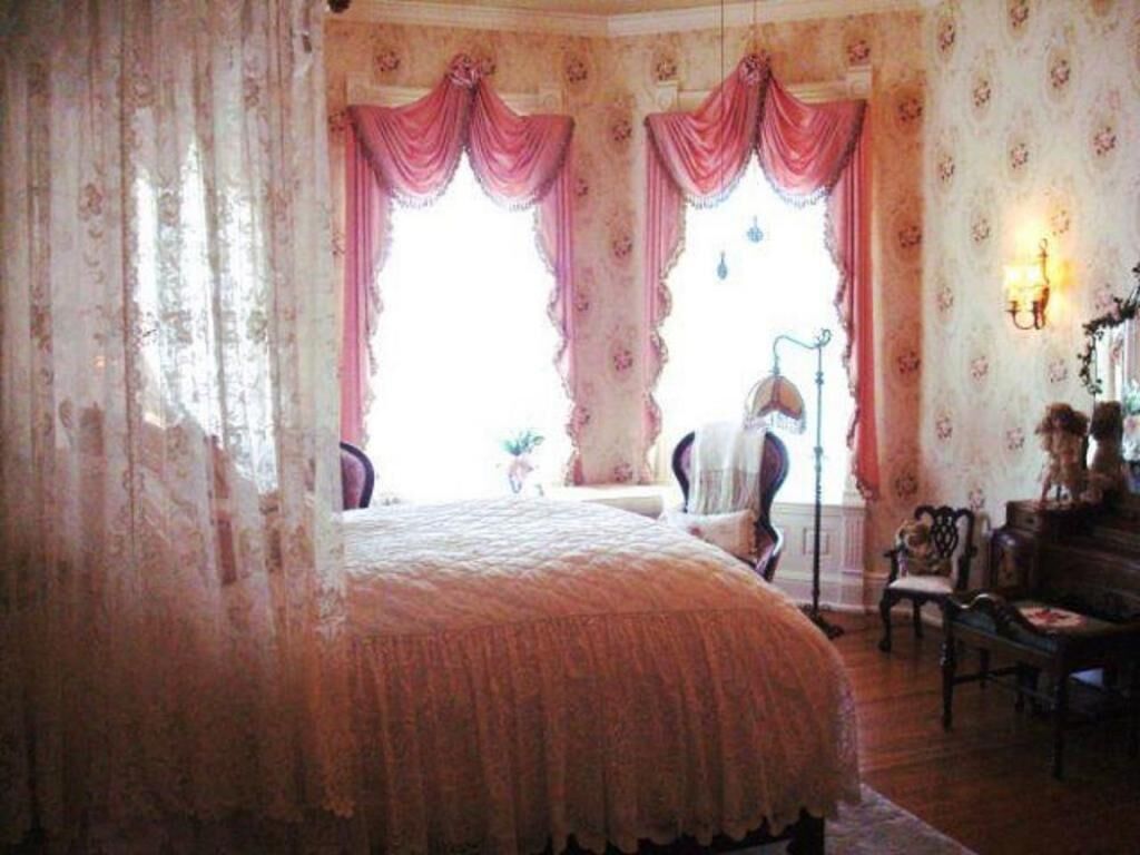 A bedroom with a large bed, two windows with pink curtains, a chair, a table, and a wall-mounted lamp.