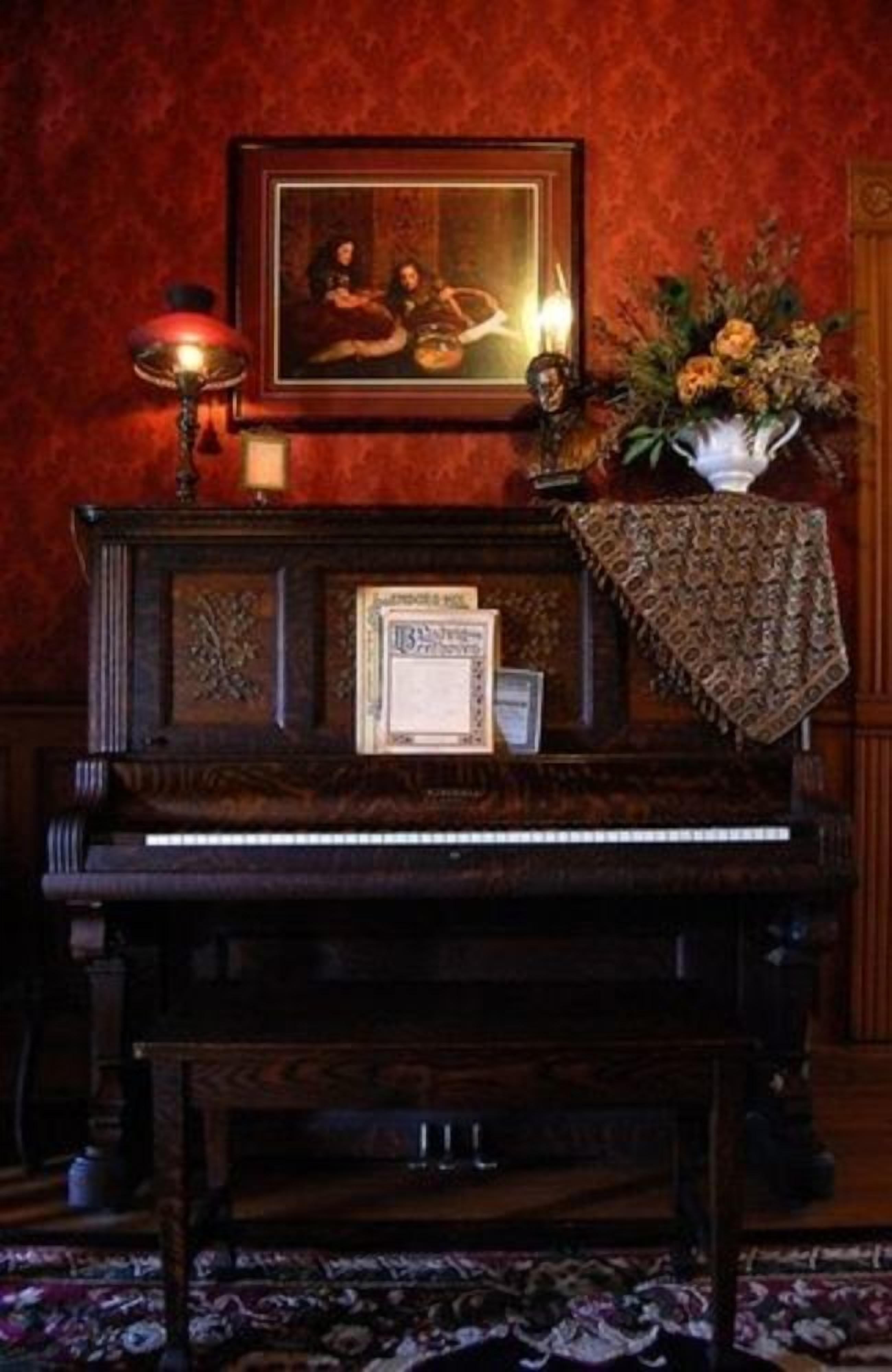 Um piano de cauda com um certificado emoldurado, uma lâmpada e um arranjo floral sobre um tapete estampado em um quarto bem iluminado.