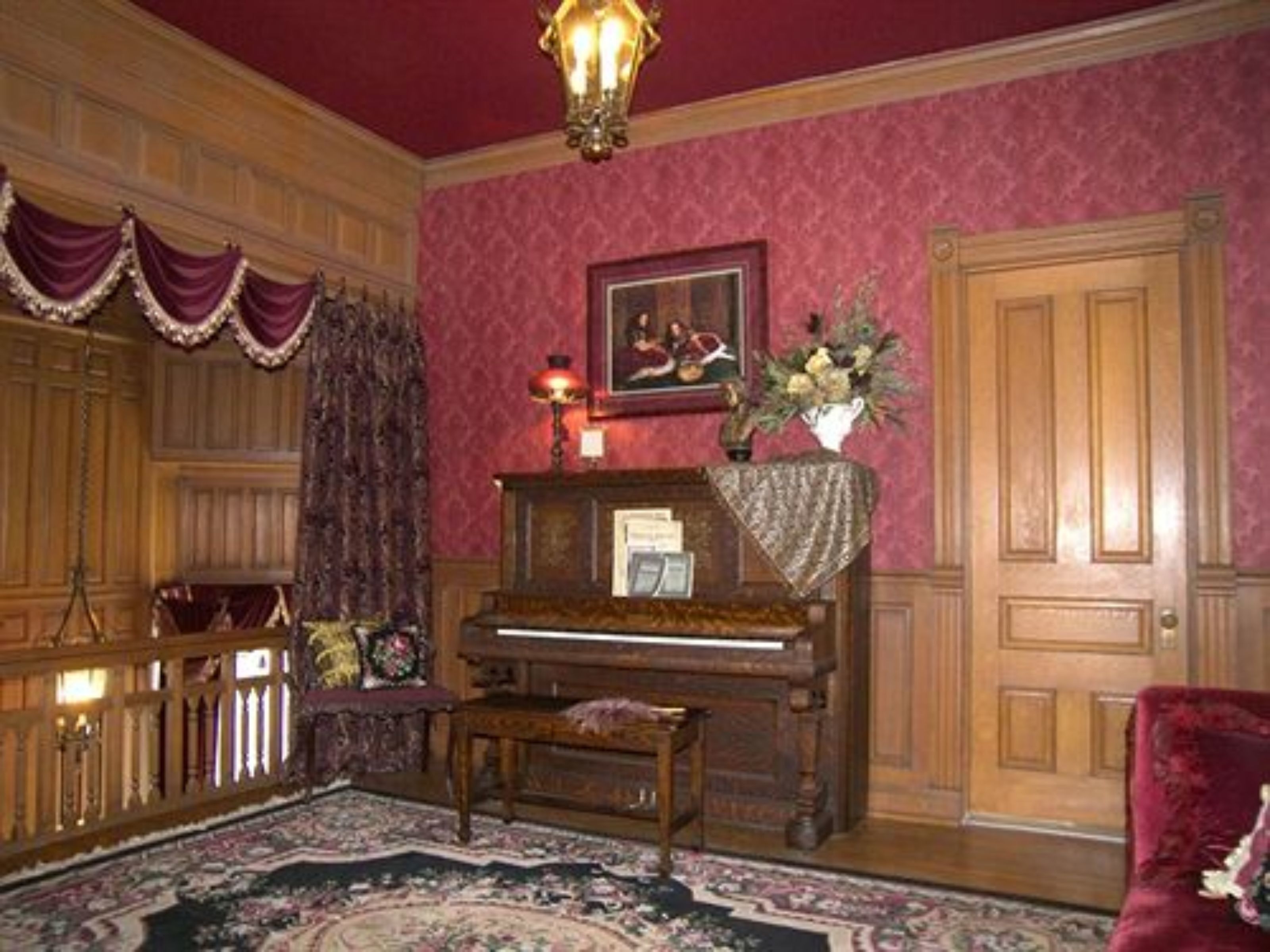 Una habitación con un piano de cola, un aparador de madera, un sillón de terciopelo rojo y una alfombra estampada.