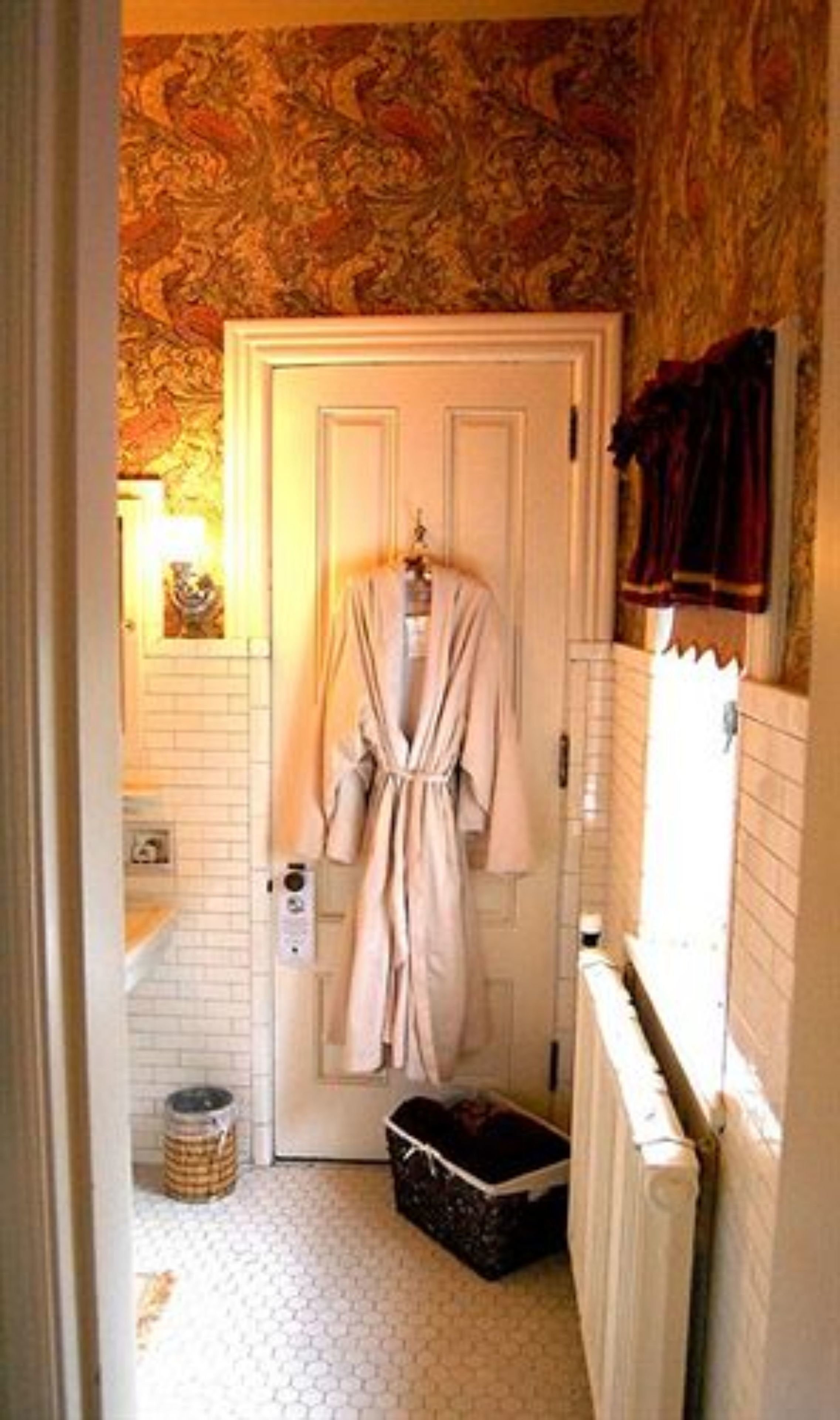 A narrow bathroom with a patterned wallpaper, a white door, and a robe hanging on the door.