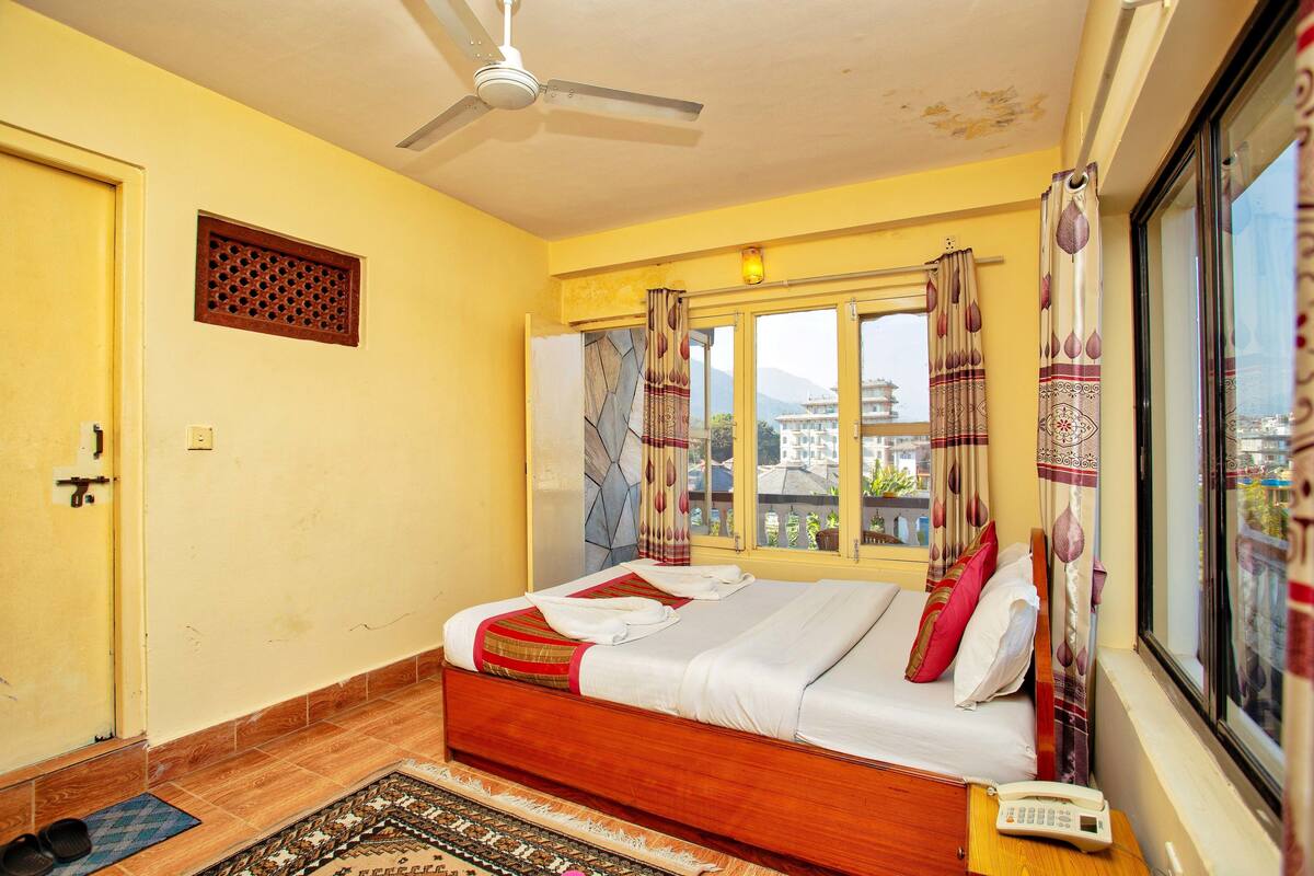A bedroom with a bed, a ceiling fan, a window with curtains, a phone on the bedside table, and a view of buildings outside.