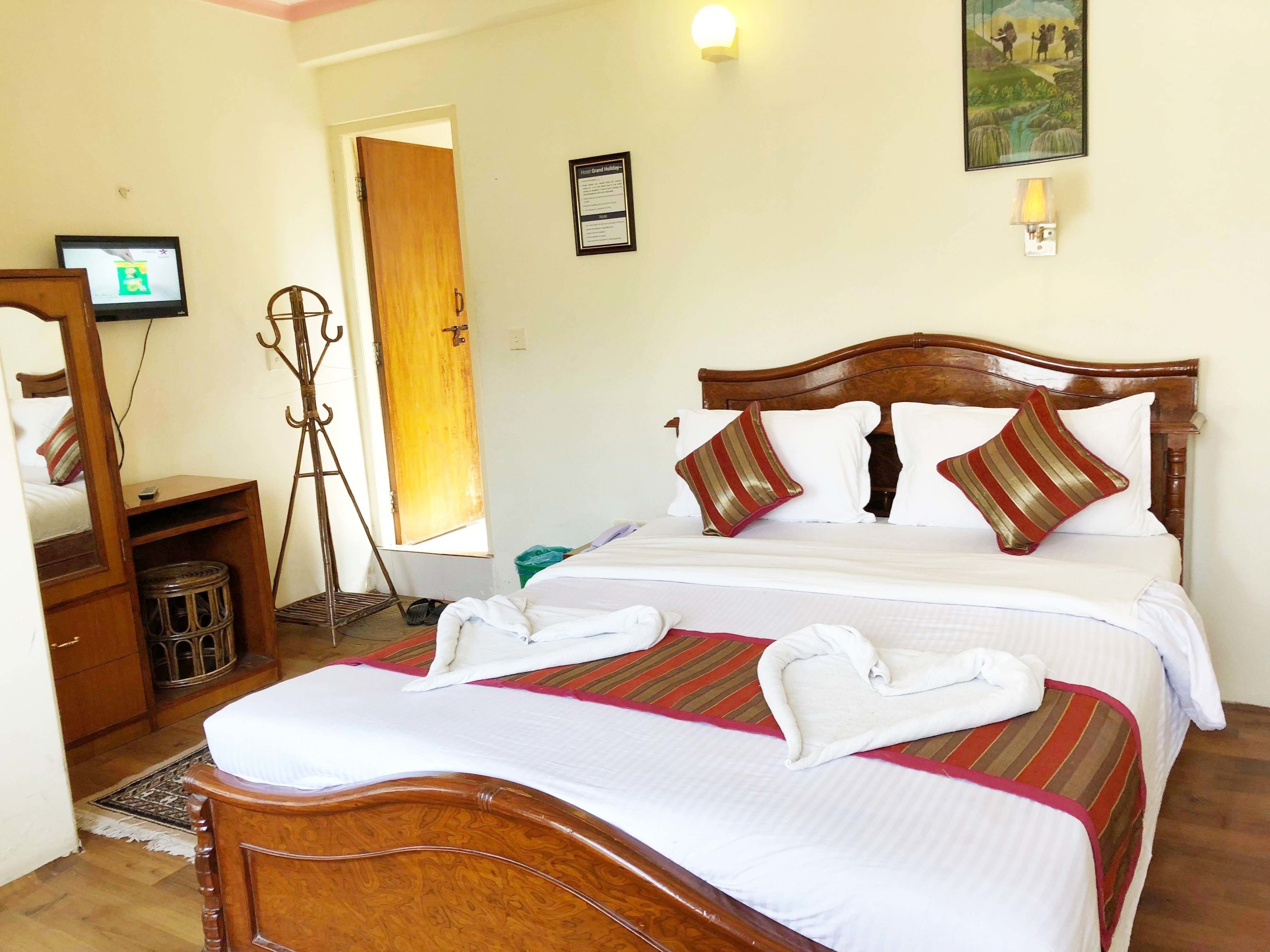 A hotel room with a wooden bed, white linens, striped pillows, a wooden nightstand, a mirror, a wall-mounted TV, and a framed picture.