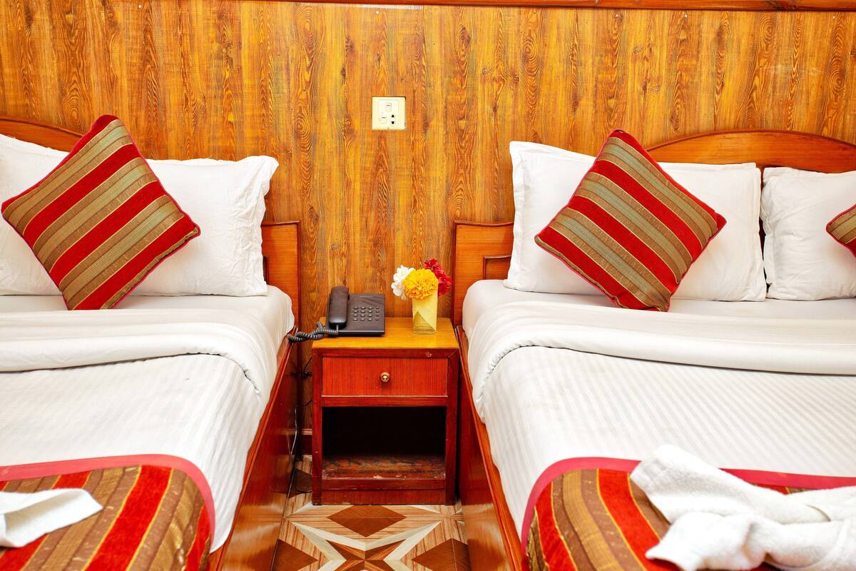 A hotel room with two beds, each with striped pillows, a bedside table with a phone and a vase of flowers, and a wooden headboard.