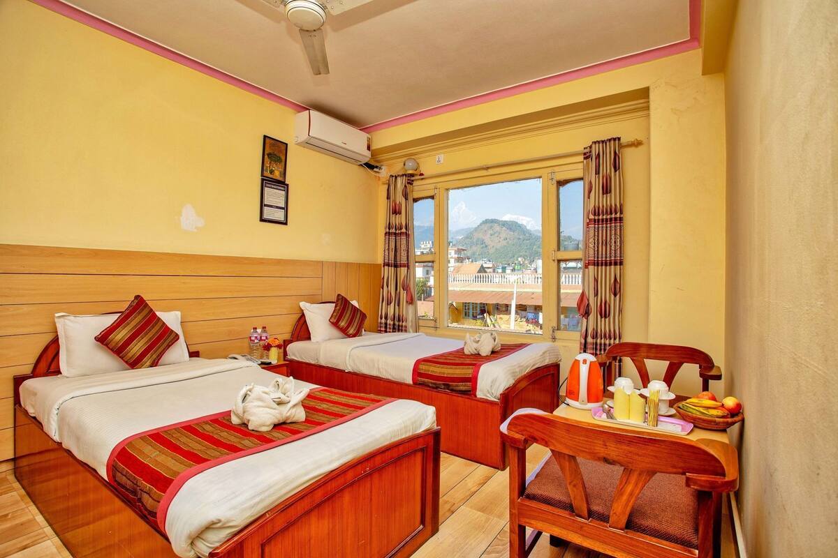 A hotel room with two beds, a wooden headboard, a window with a mountain view, and a table with a kettle and fruit.