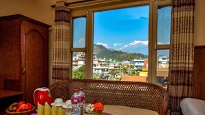A room with a large window, wicker furniture, a table with fruits and bottles, and a view of a mountainous landscape. - Hotel Grand Holiday (Pokhara)