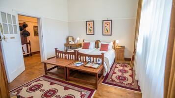 A hotel room with a double bed, a wooden bench, a small table, two bedside tables, a nightstand, and a patterned rug.