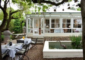A covered outdoor dining area with tables and chairs, a small pond with a bird, and a white building with large windows. - Bartholomeus Klip Farmhouse (Hermon)