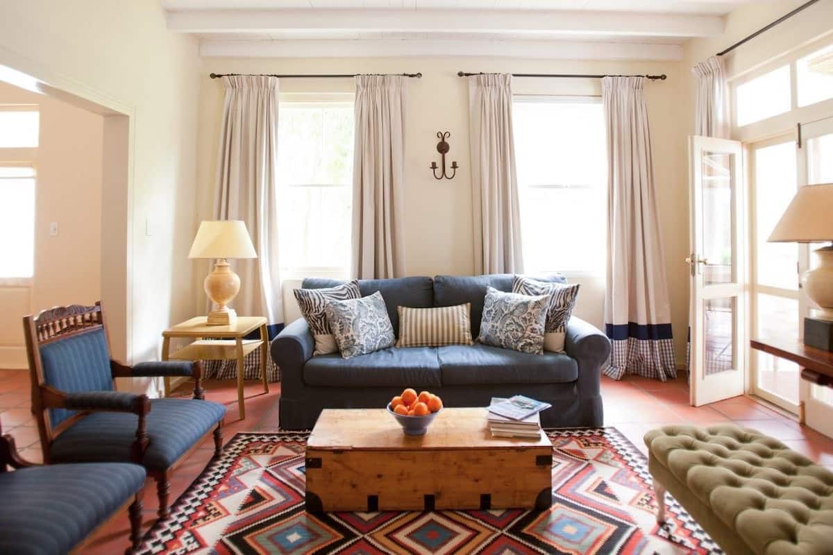 Una sala de estar con sofá, sillones, una mesa de centro con frutero, alfombras estampadas y cortinas.