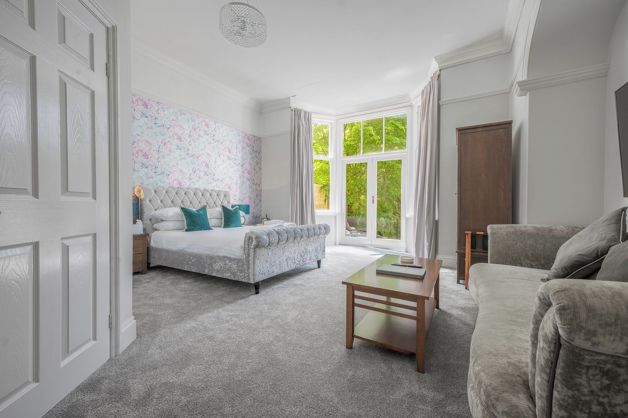 A bedroom with a bed, a sofa, a wooden table, and a large window with a view of greenery.