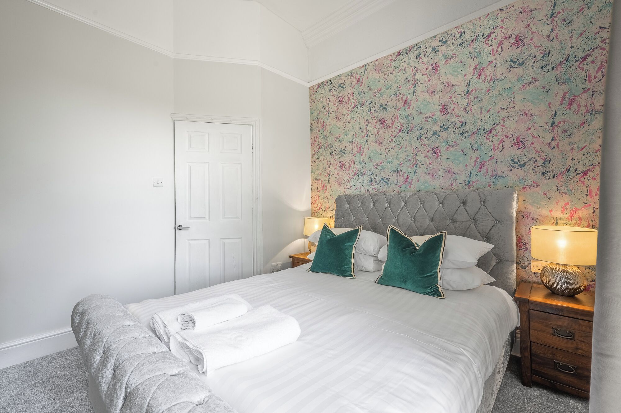 Een net opgemaakt bed met witte lakens en twee groene kussens, een gestuufd hoofdeinde en een wand met een bloemenpatroon.
