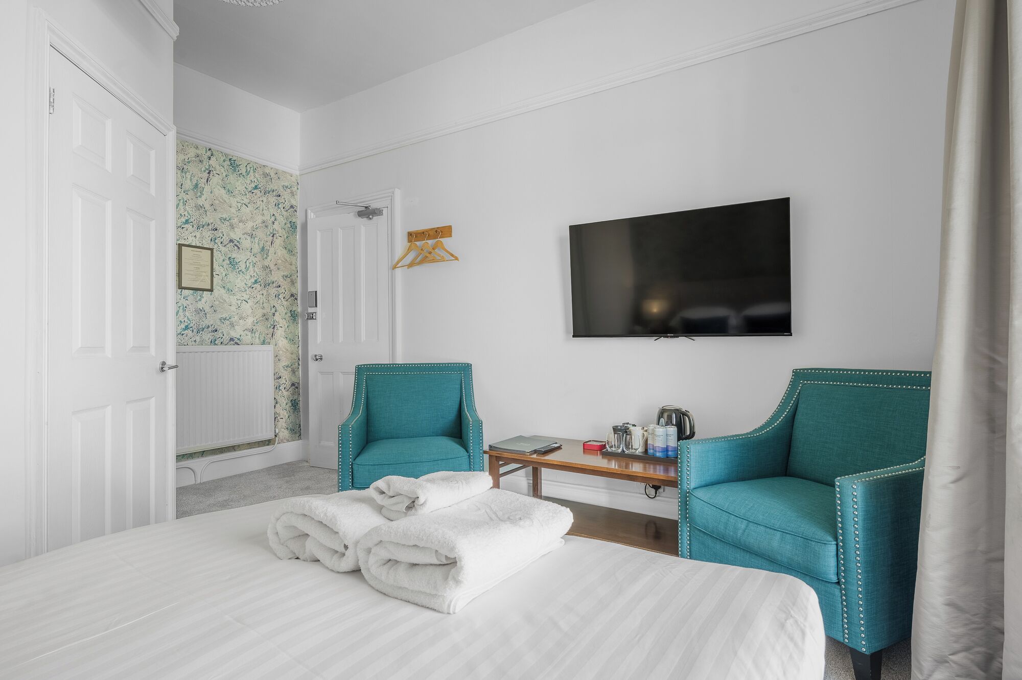 A hotel room with a bed, two teal armchairs, a small table with a kettle and cups, a TV on the wall, and a door leading to a bathroom.