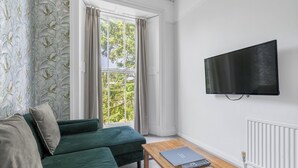 En stue med en grøn sofa, et træ sofabord, et fladskærms-tv og et vindue med gardiner.