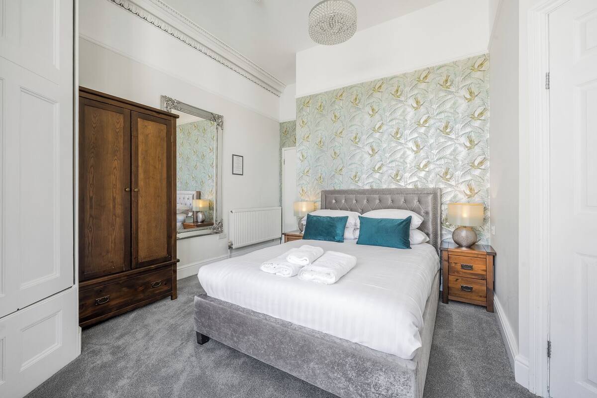 A bedroom with a large bed, a wardrobe, a mirror, and a patterned wallpaper.