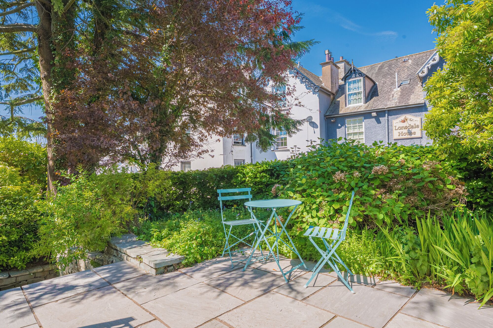 Ein Garten mit einem Kiesweg, zwei blauen Stühlen und einem Gebäude mit dem Schild "Lonsdale House".