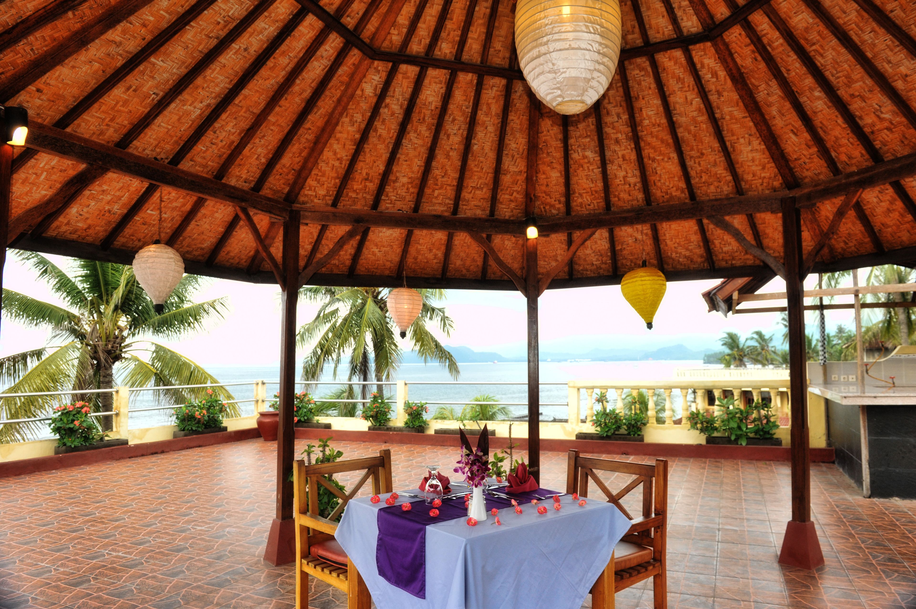 A covered outdoor dining area with a table set for two, overlooking a scenic view of the sea and palm trees.
