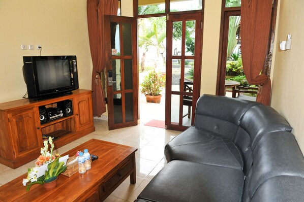 A living room with a leather sofa, a wooden coffee table with a flower arrangement, a television on a wooden cabinet, and a view of a garden through glass doors.