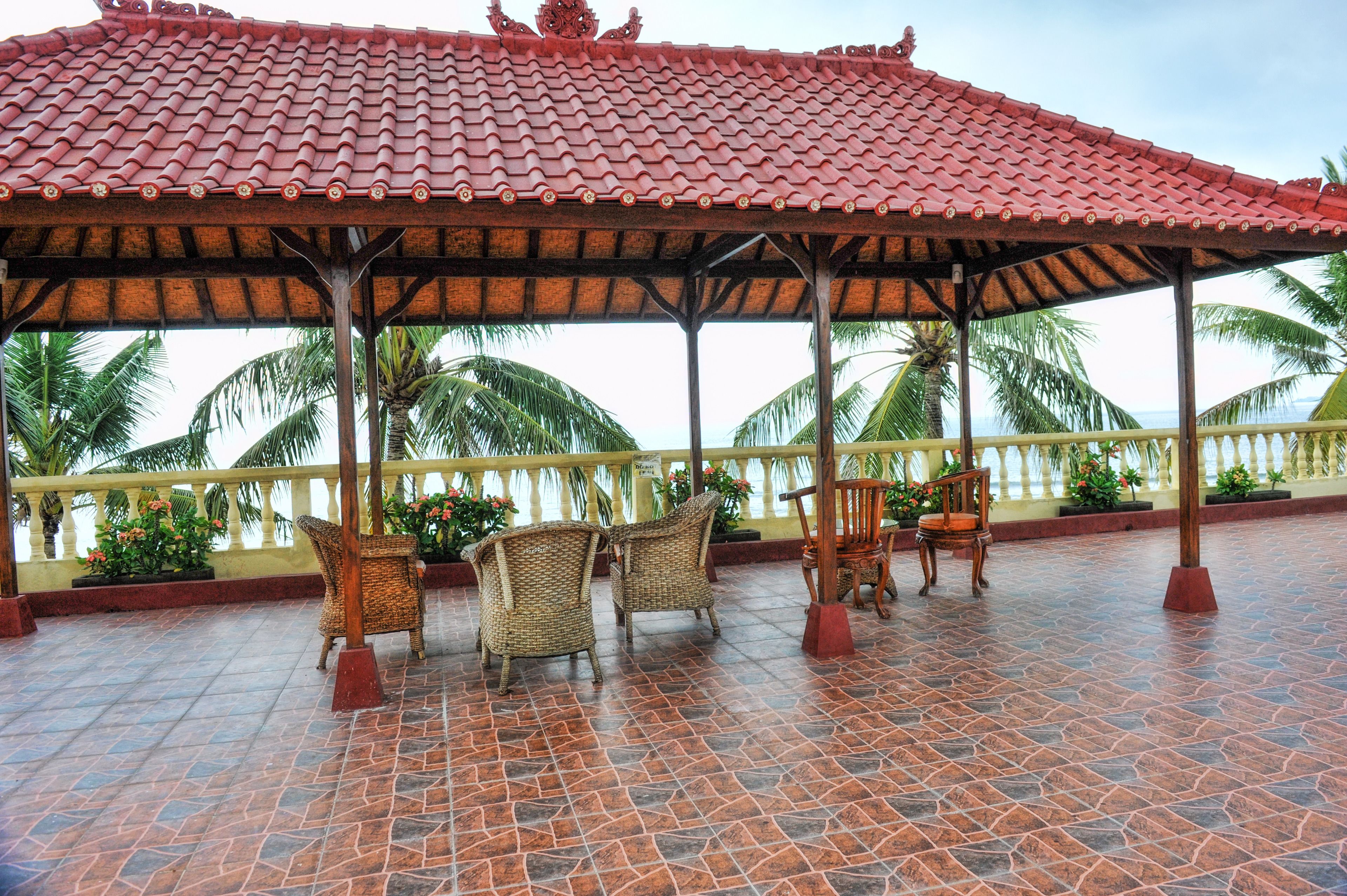 A covered outdoor seating area with wicker chairs and a wooden chair, a tiled floor, and a view of palm trees and the ocean.
