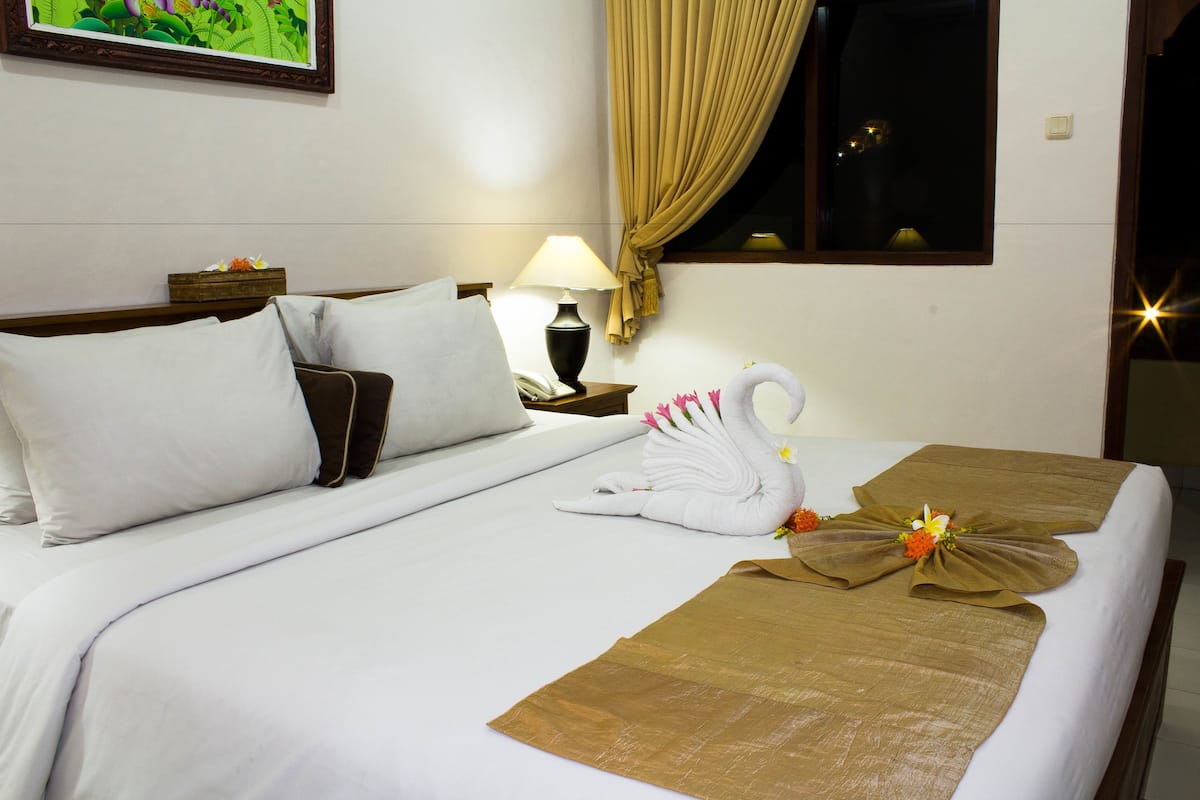A hotel room with a neatly made bed, a bedside lamp, a window with curtains, and a framed picture on the wall.