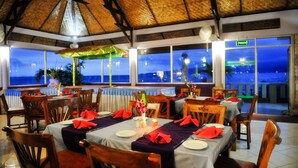 A restaurant with tables set for dining, a thatched roof, and a view of the sea. - Bali Palms Resort (Manggis)