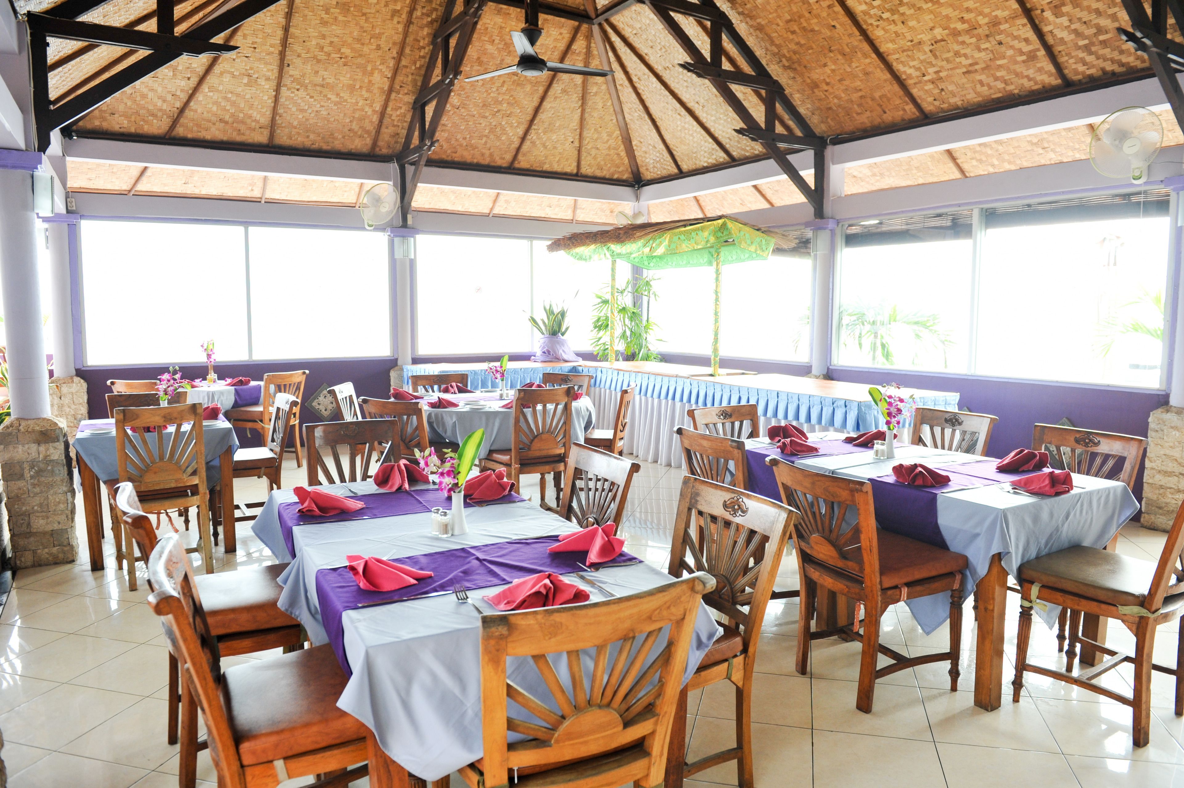 Un restaurante con mesas y sillas de madera, cubierto por un techo de paja, y ventanas amplias que dejan entrar luz natural.